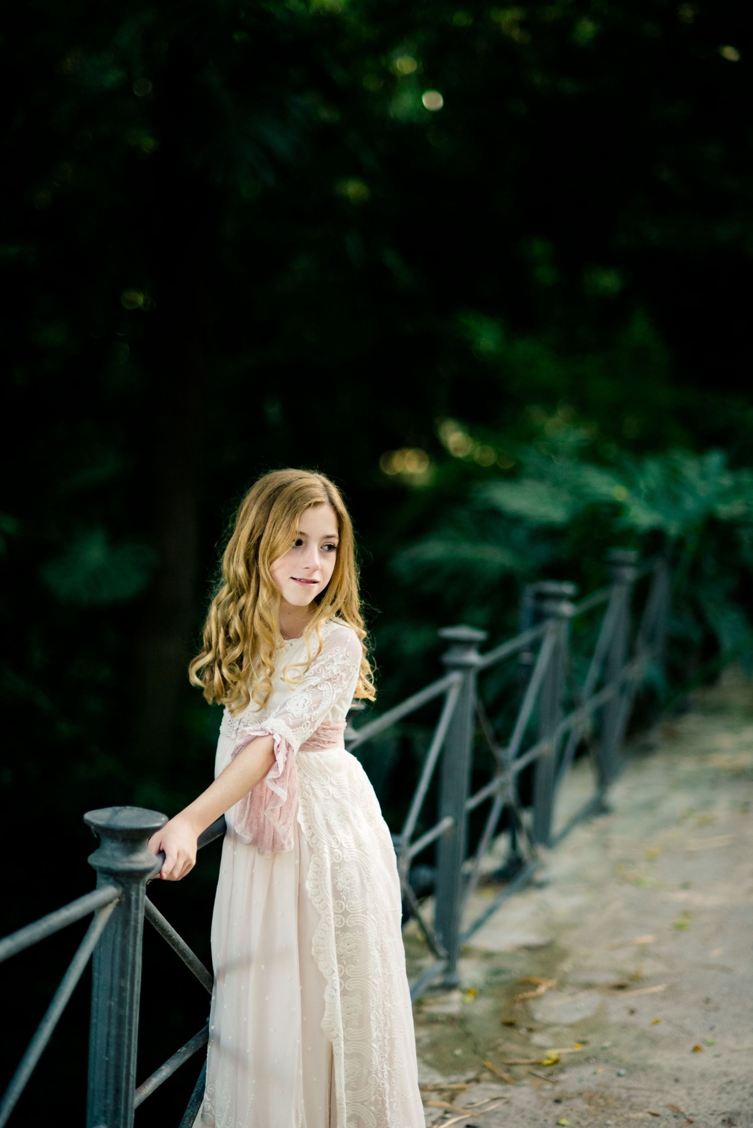 Blonde girl in pink lace dress smiles while leaning on iron fence in garden setting.