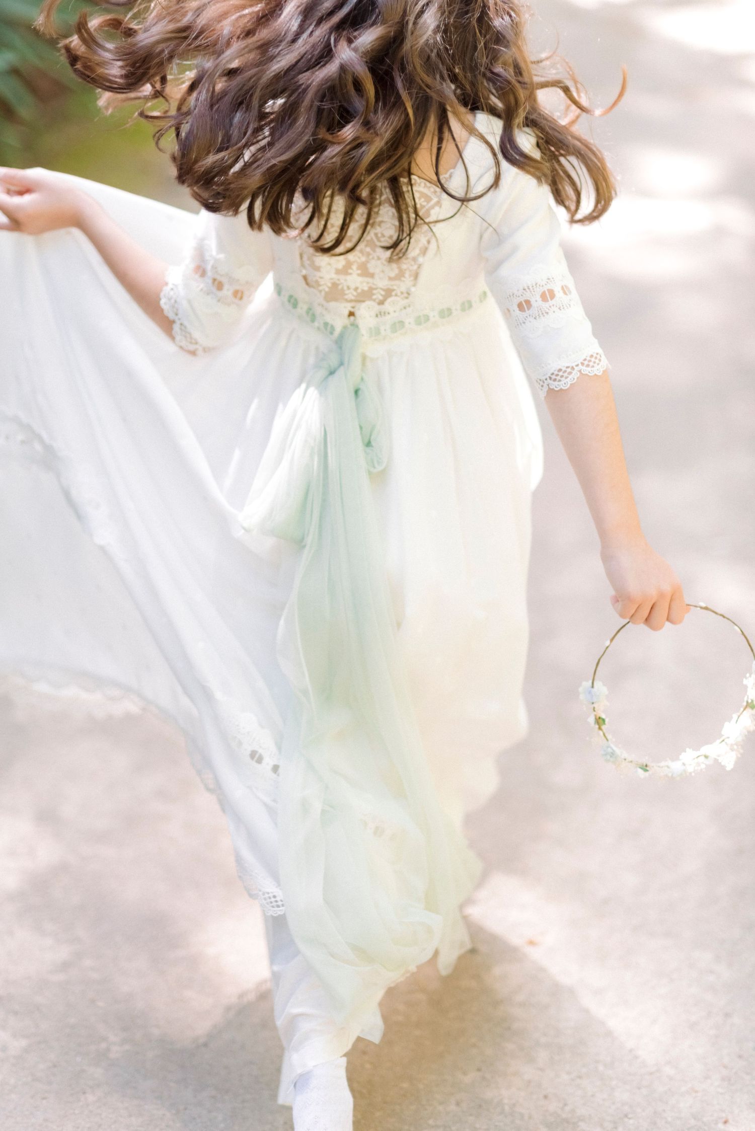 A girl in a flowing white and mint dress twirls gracefully while holding a floral crown outdoors.