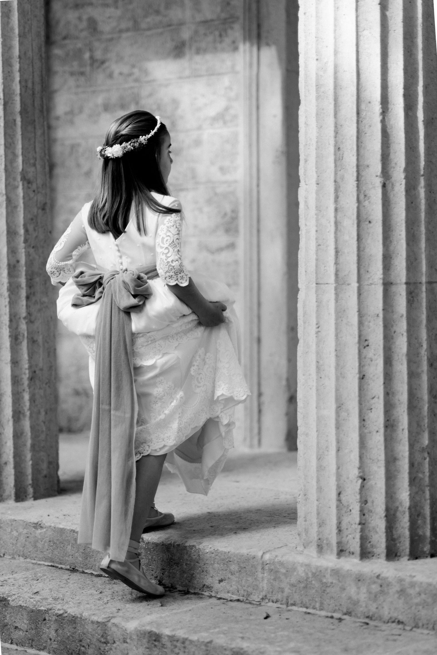 Girl in white lace communion dress and floral crown stands between stone columns in black and white photo.