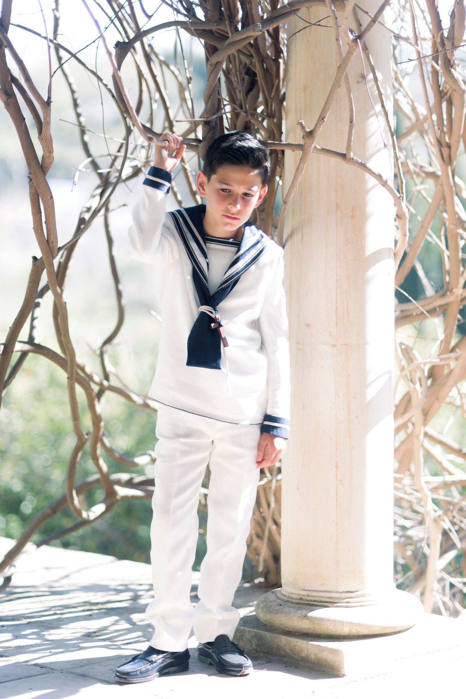 Boy in white navy sailor suit leans against stone column among bare branches outdoors.