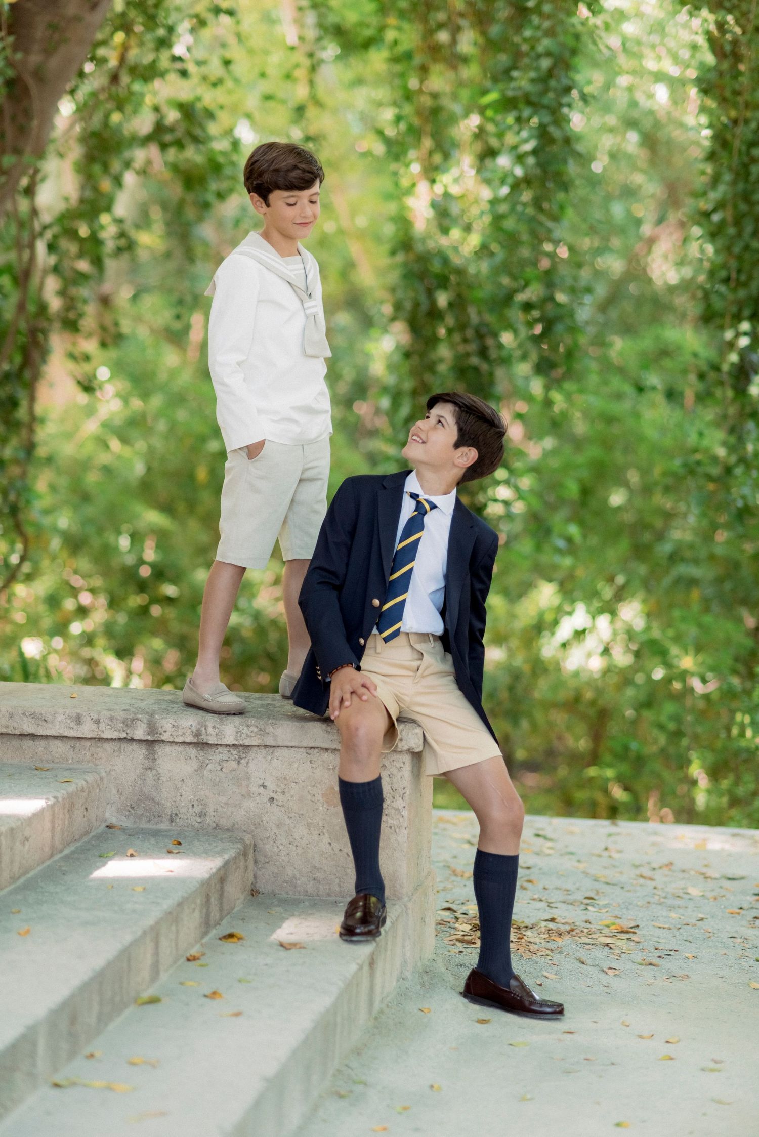 Two boys in formal attire on stone steps, one in navy blazer with tie, one in white.