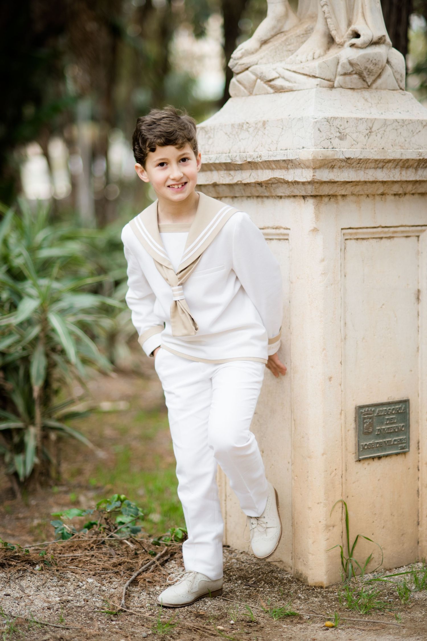 Smiling boy in white sailor suit leaning on stone statue.