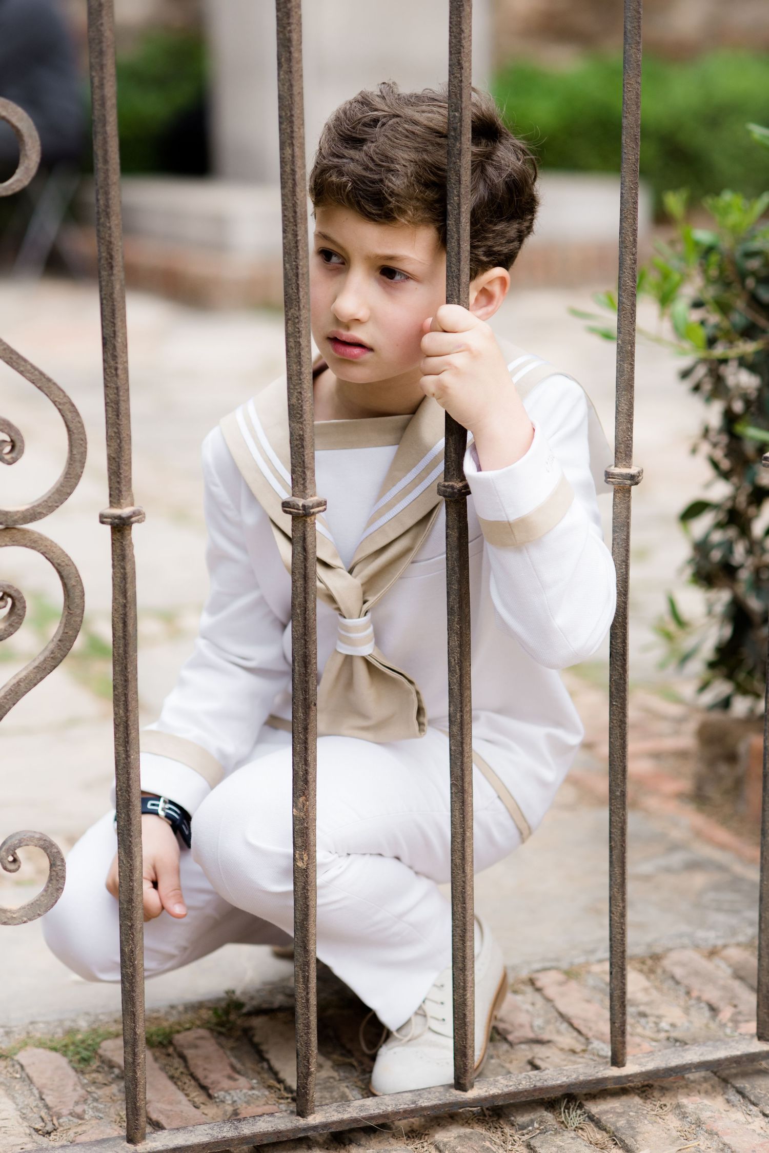 A boy in a white sailor communion outfit crouches behind decorative iron bars outdoors.