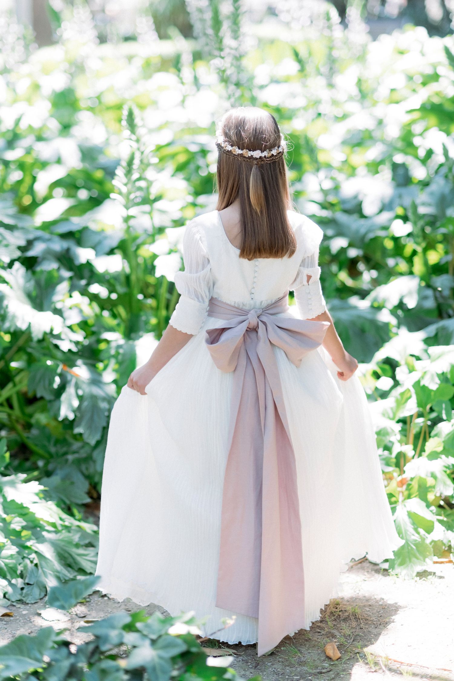 Girl in white communion dress with large bow and floral crown viewed from behind in garden.
