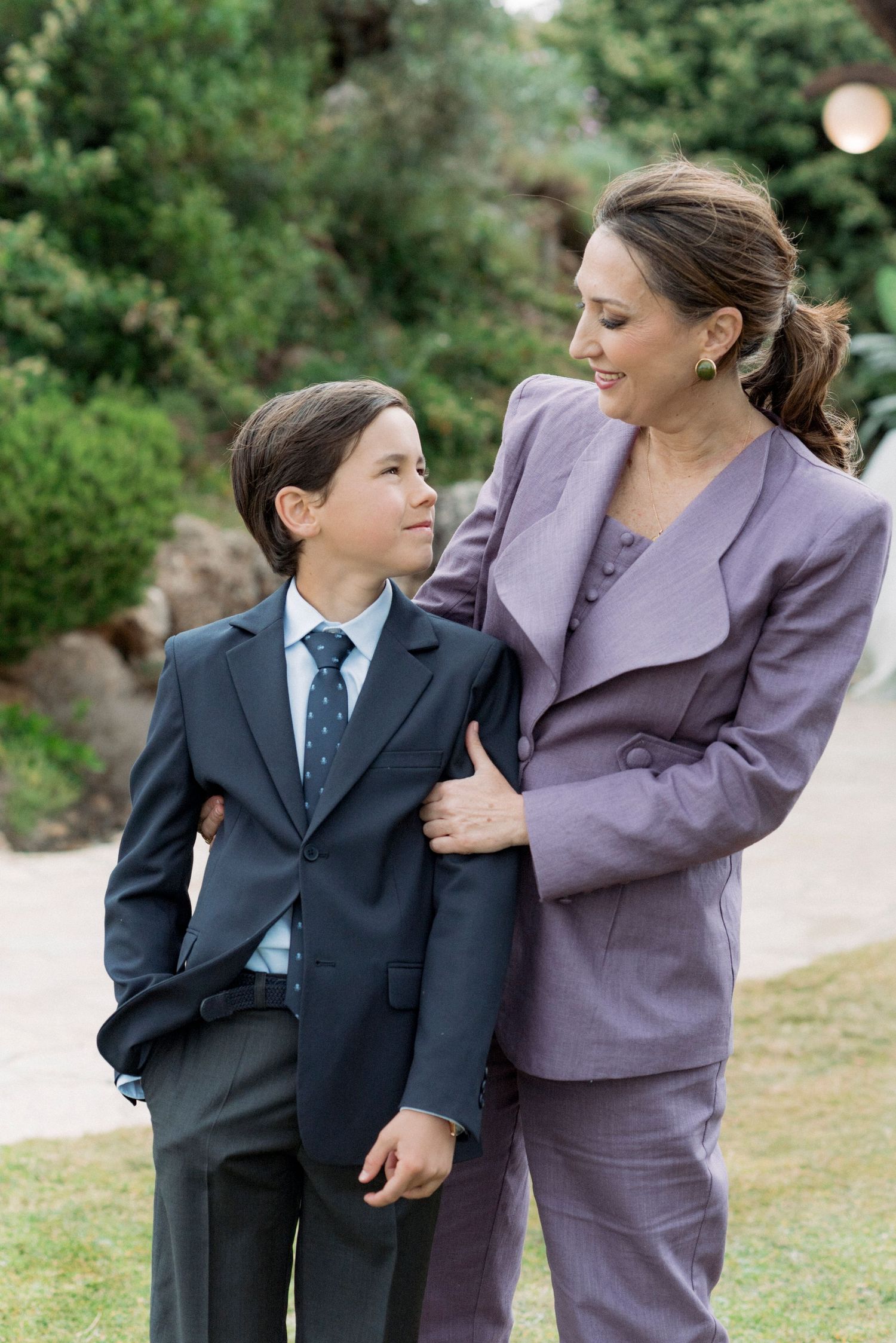 Boy in navy suit with tie smiling at elegantly dressed woman in lavender pantsuit outdoors.