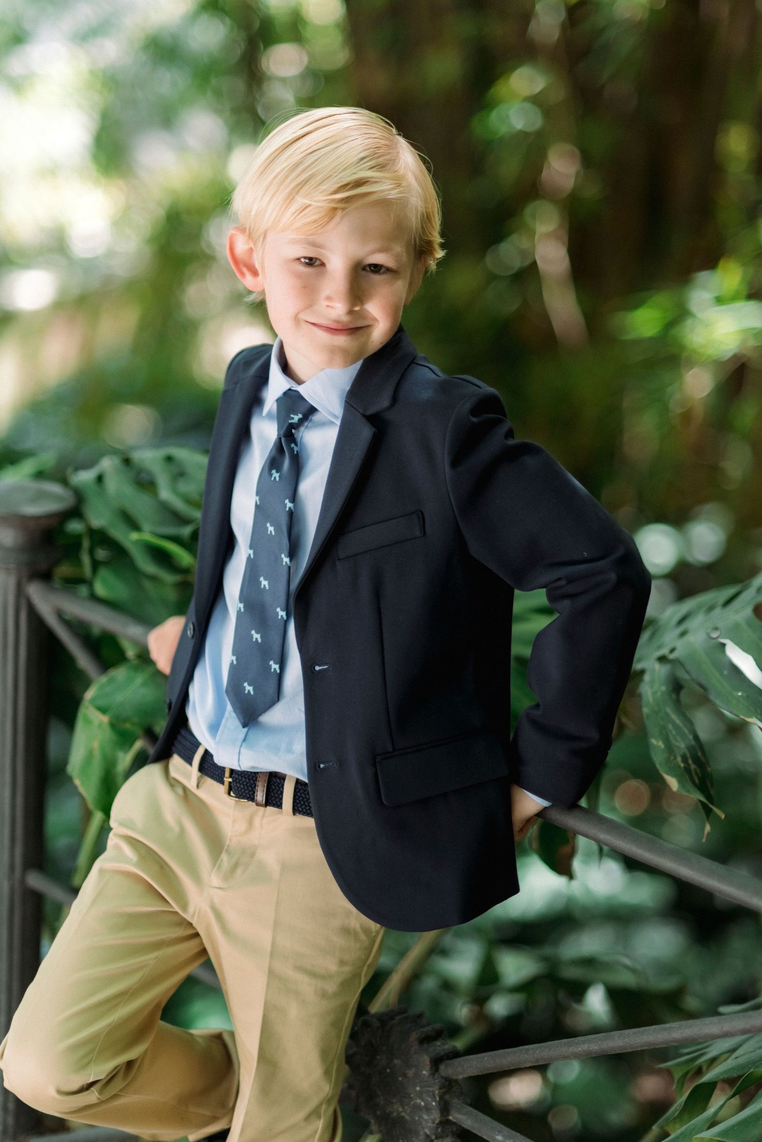 Smiling blonde boy in navy blazer, tie, and khaki pants poses confidently in lush garden setting.