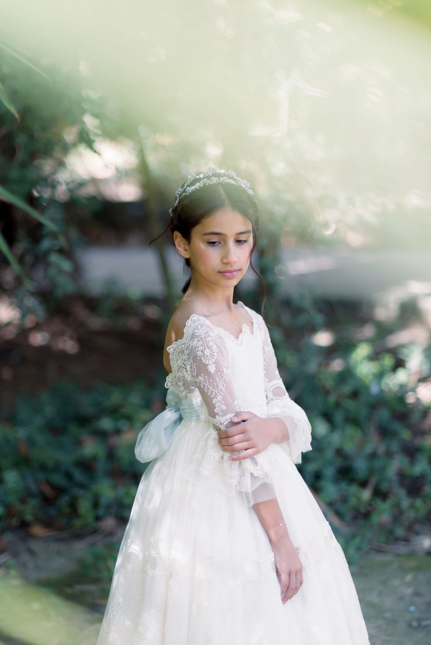 Girl in elegant white lace First Communion gown with floral crown in garden setting.