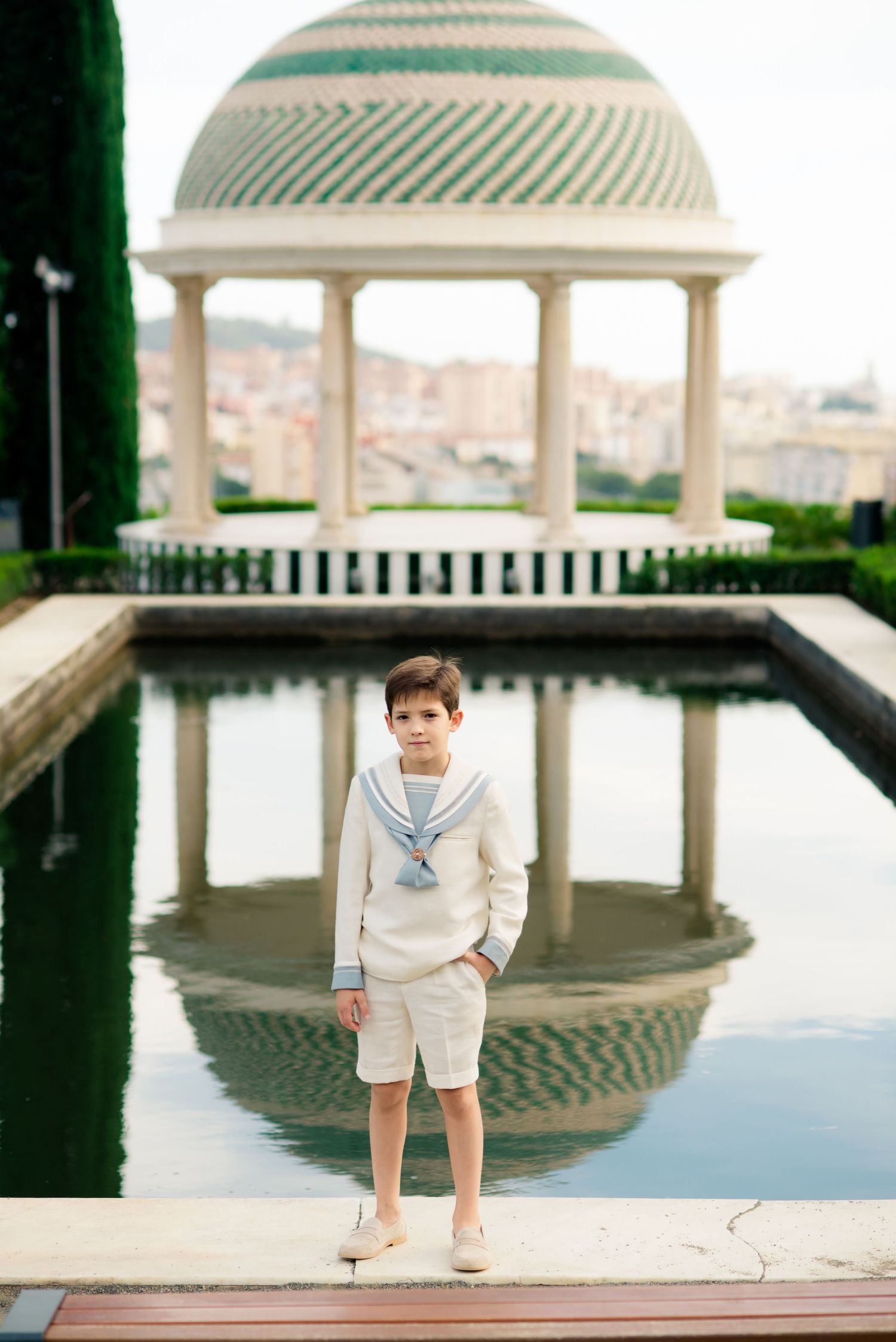 Young boy in cream sailor suit stands beside reflecting pool with domed pavilion backdrop.
