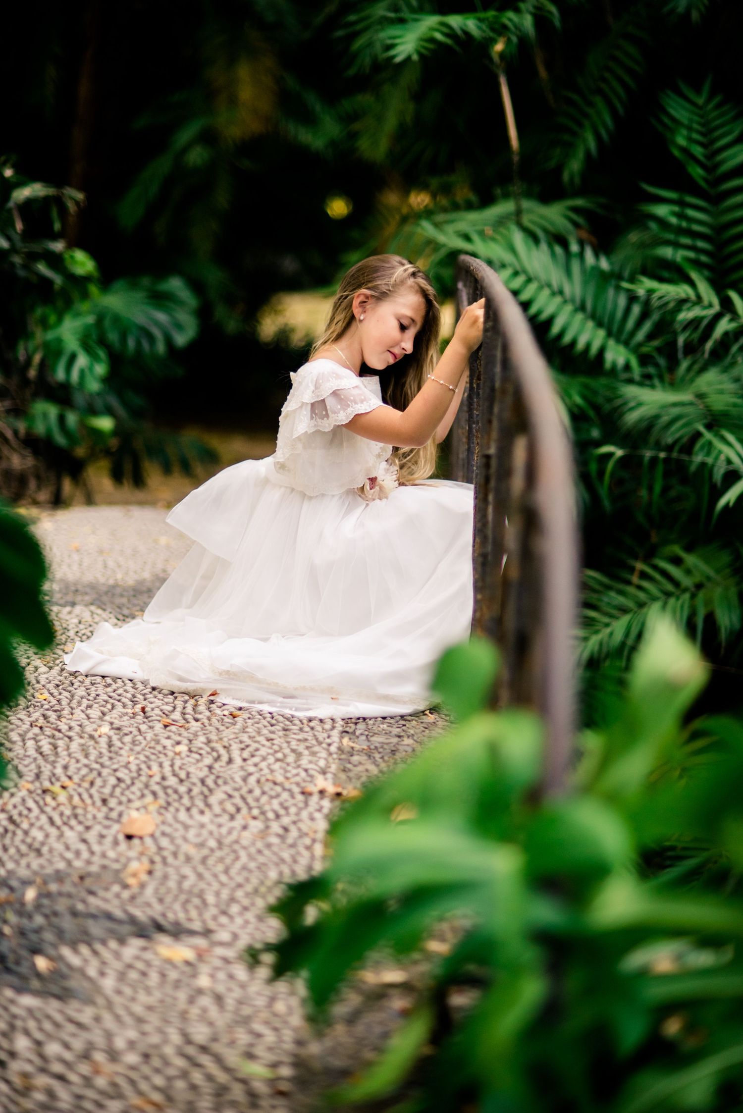 Young girl in white tulle dress leans on gate surrounded by tropical garden greenery.