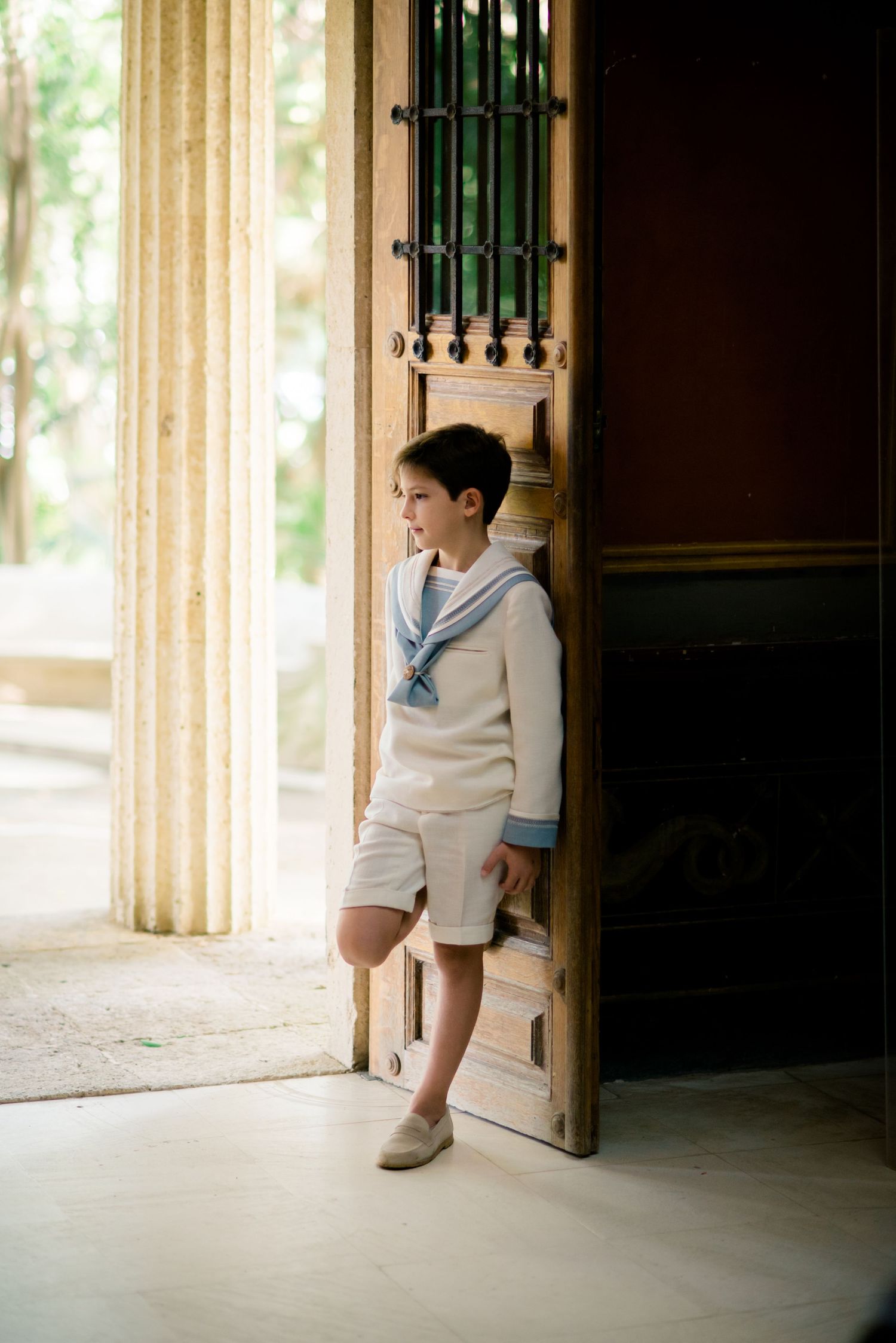 Boy in cream sailor outfit leans against ornate wooden door in elegant architectural setting.