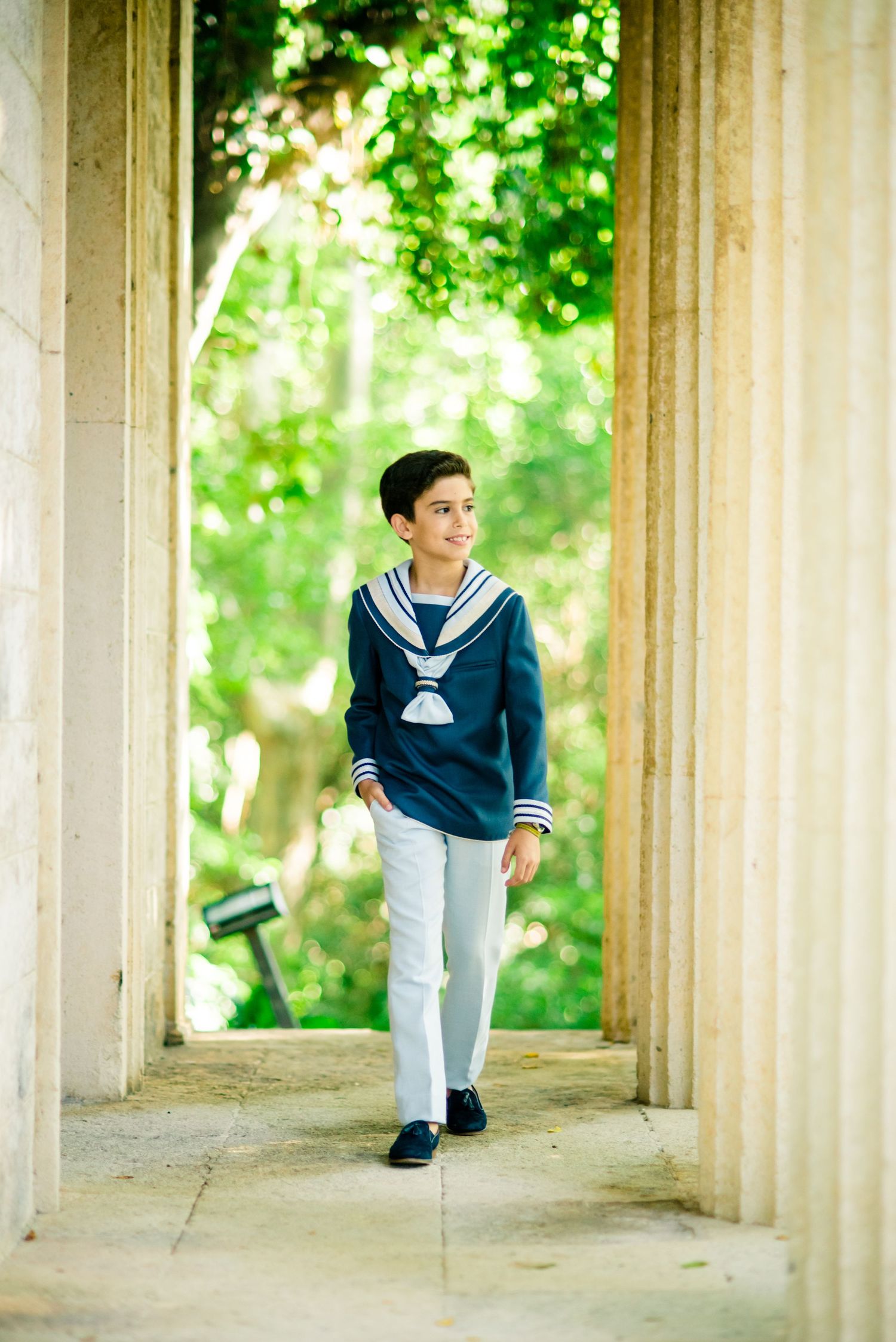 Boy in navy sailor sweater and white pants walking through stone colonnade in garden.