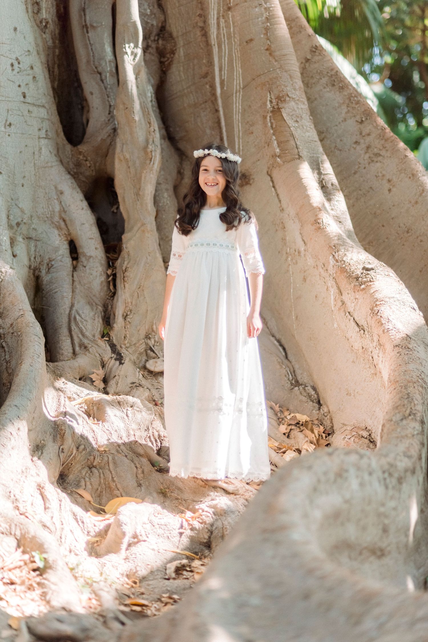 A smiling girl in a white communion dress and floral crown stands before a large ancient tree.