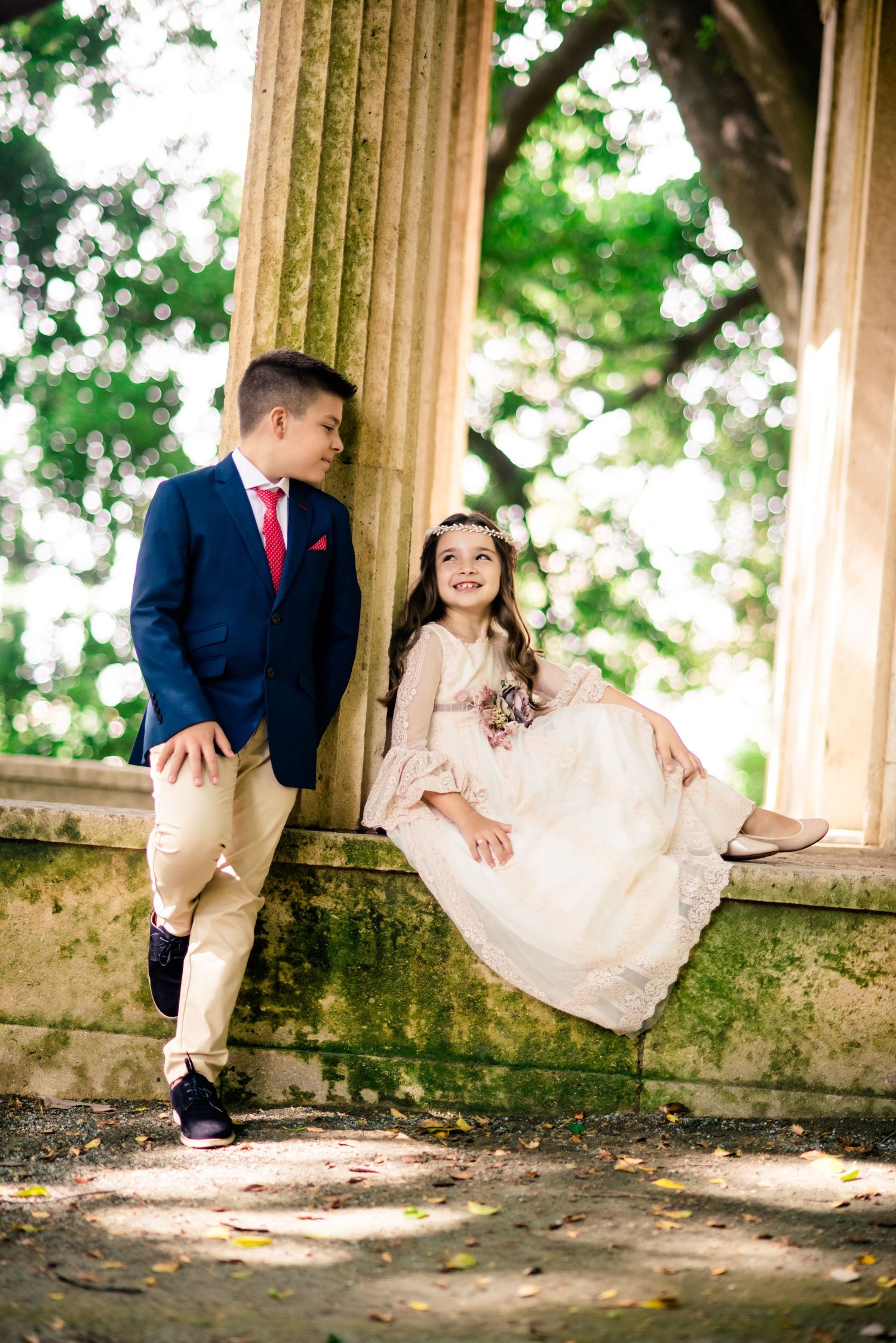 Boy in navy suit and girl in white communion dress posing together at stone columns.