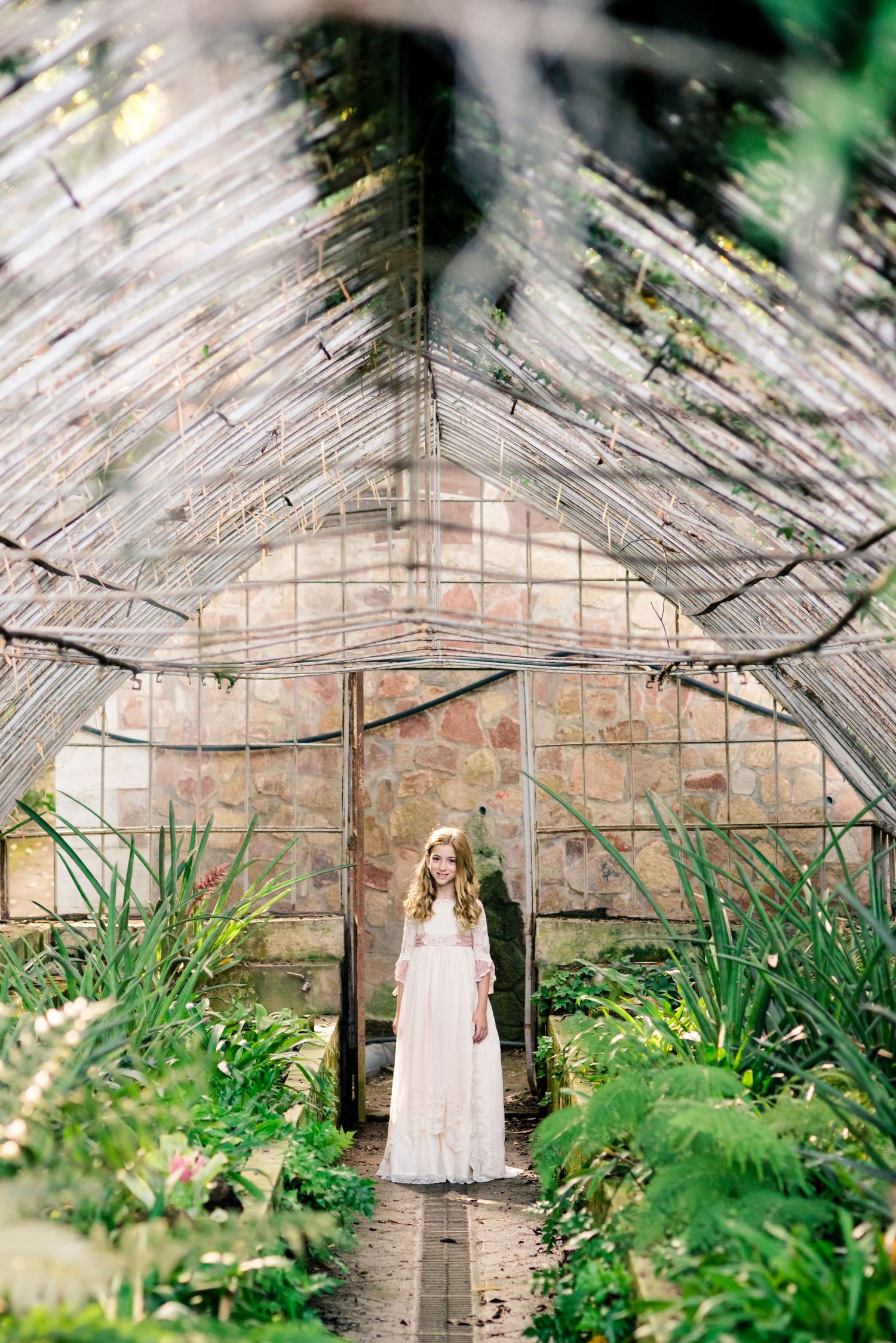 Young girl in white lace communion dress stands in lush vintage greenhouse interior.