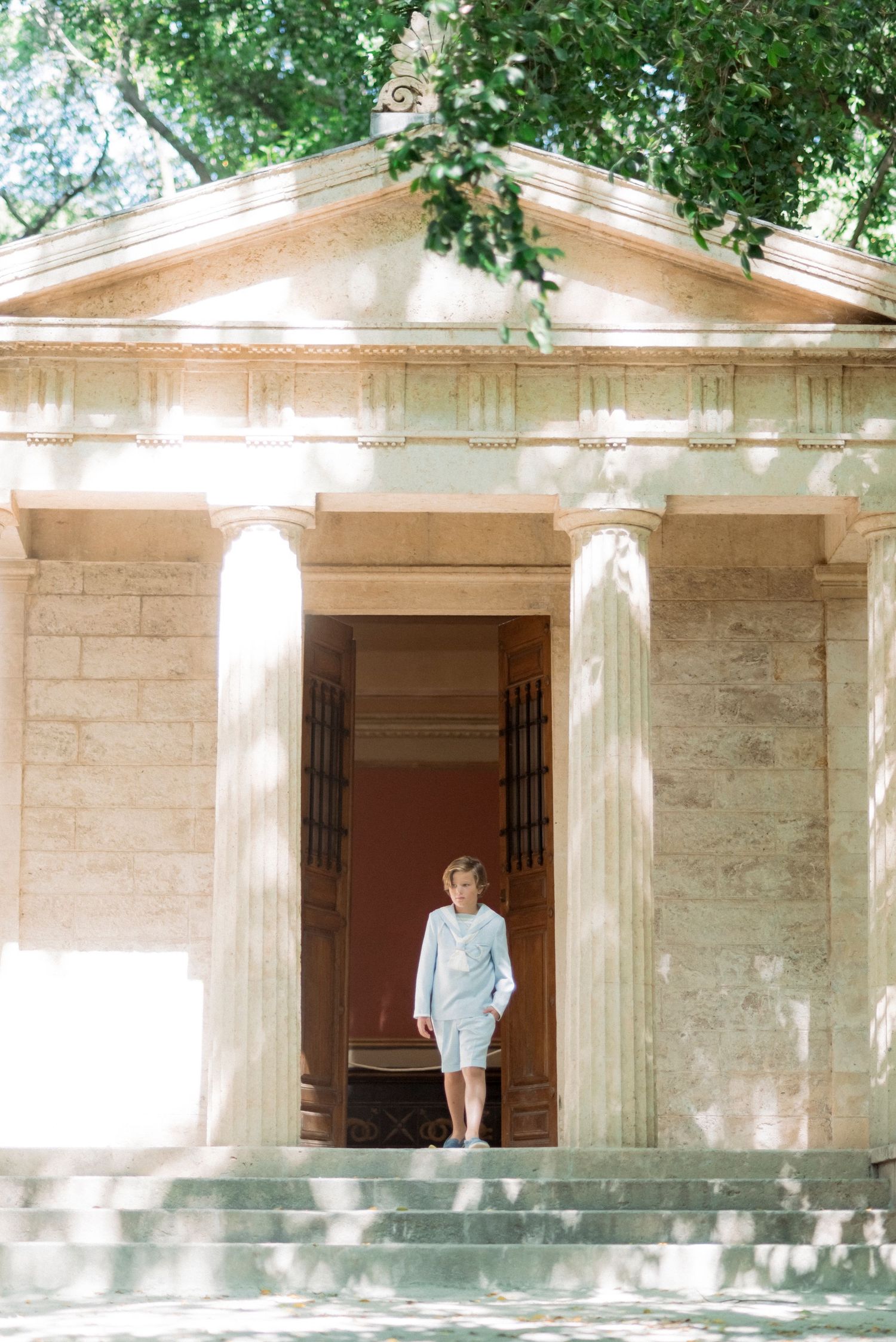 Boy in light blue suit standing between classical columns of a neoclassical building entrance.
