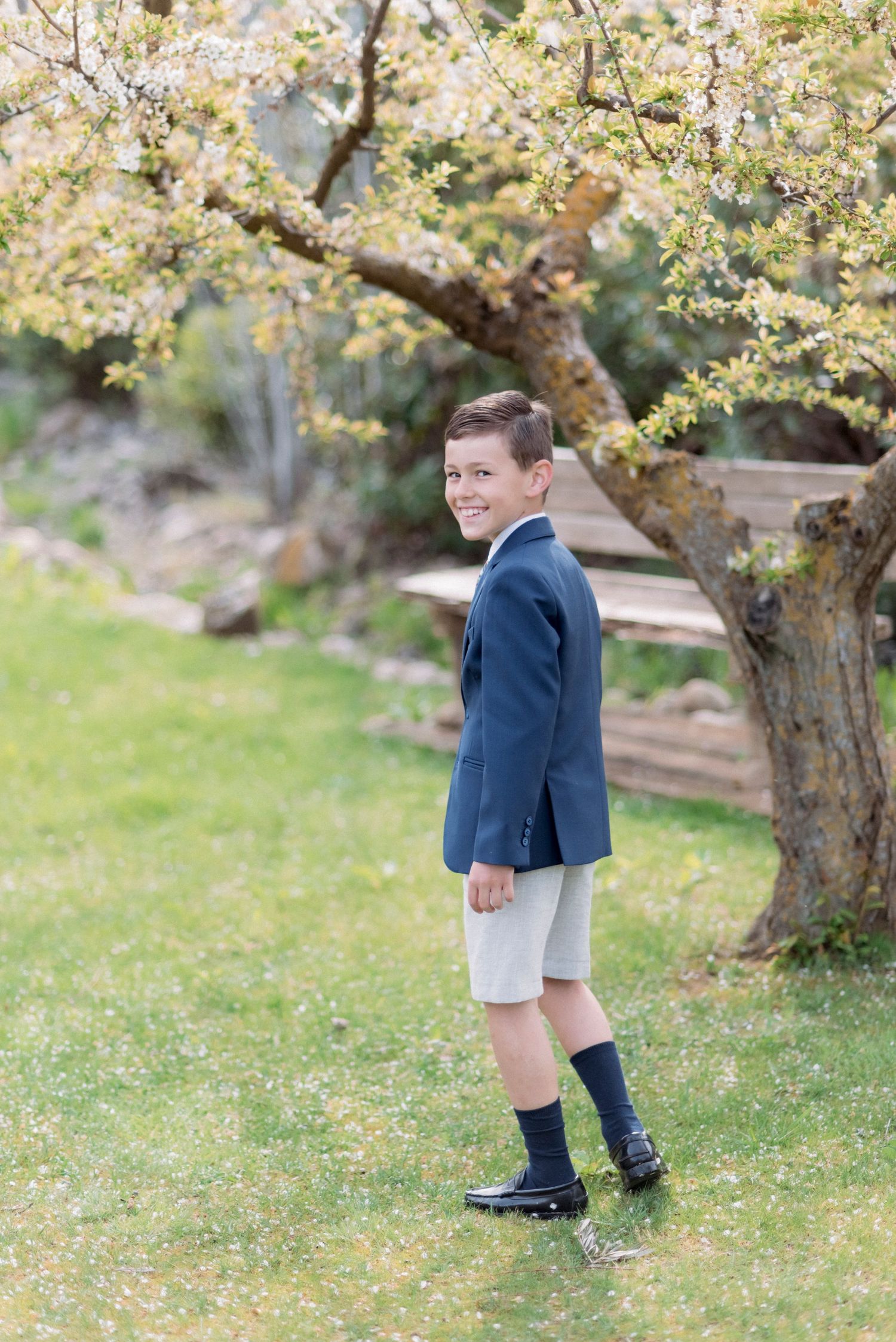 A boy in a navy blazer and shorts smiles while standing on a green lawn beneath a blossoming tree.