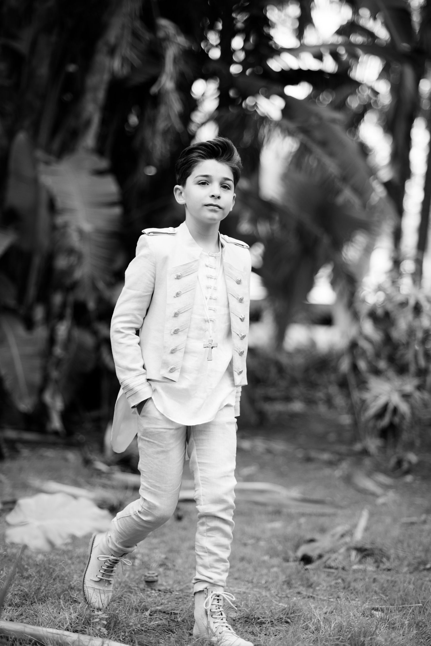 A boy in a white military-style jacket poses outdoors in a black and white photo.