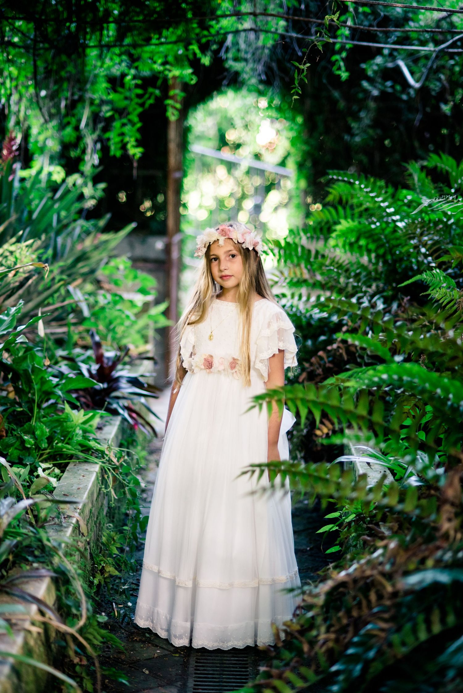 Young girl in white dress and floral crown stands in lush green garden greenhouse.