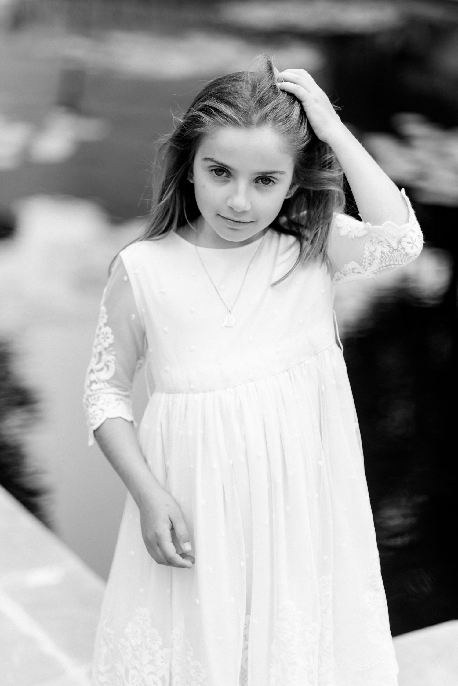 Young girl in white lace dress touches her hair in elegant black and white portrait photo.