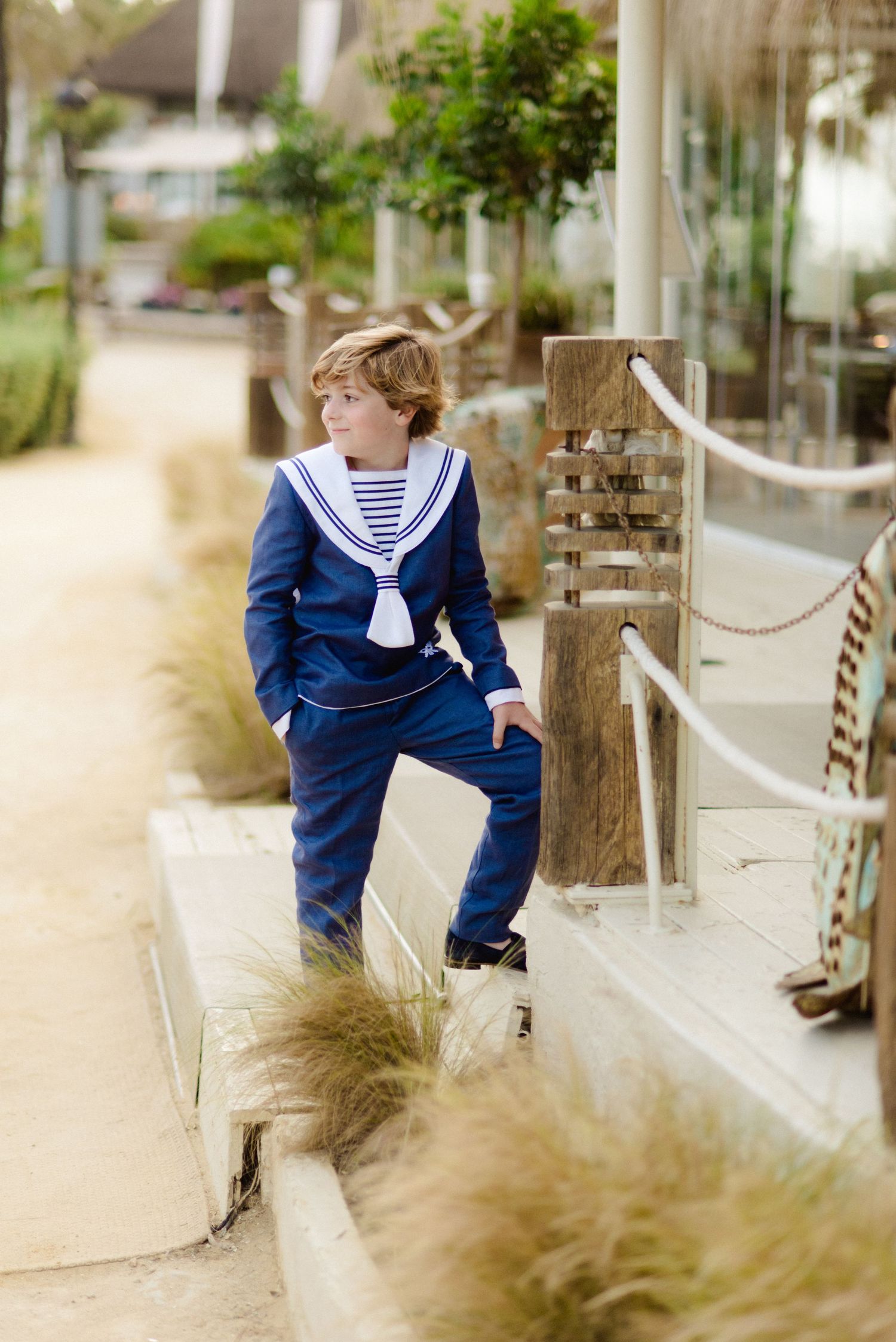 Boy in navy sailor suit posing on steps near a nautical rope railing outdoors.