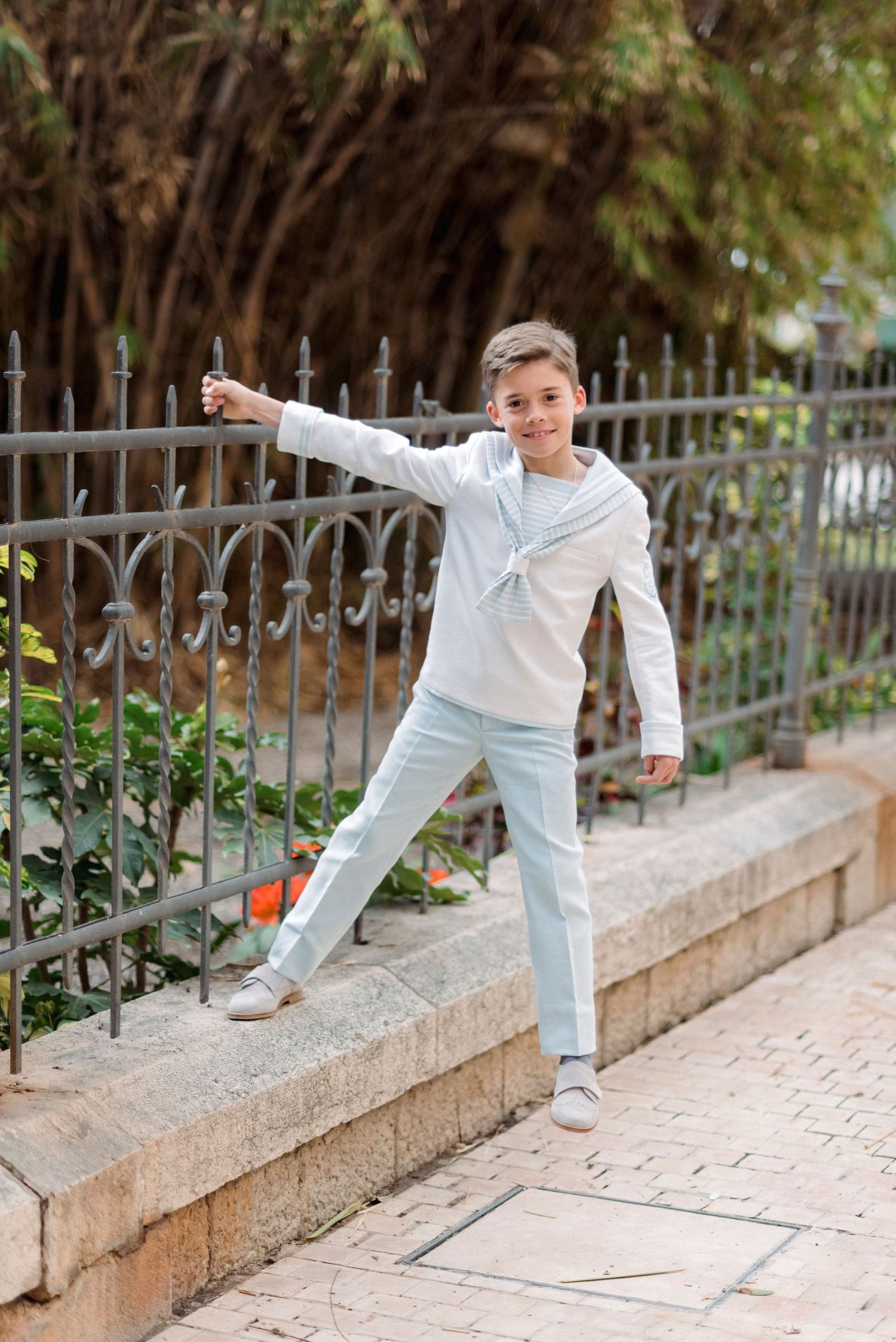 Boy in white hoodie and light blue pants stands by ornate iron fence in outdoor setting.
