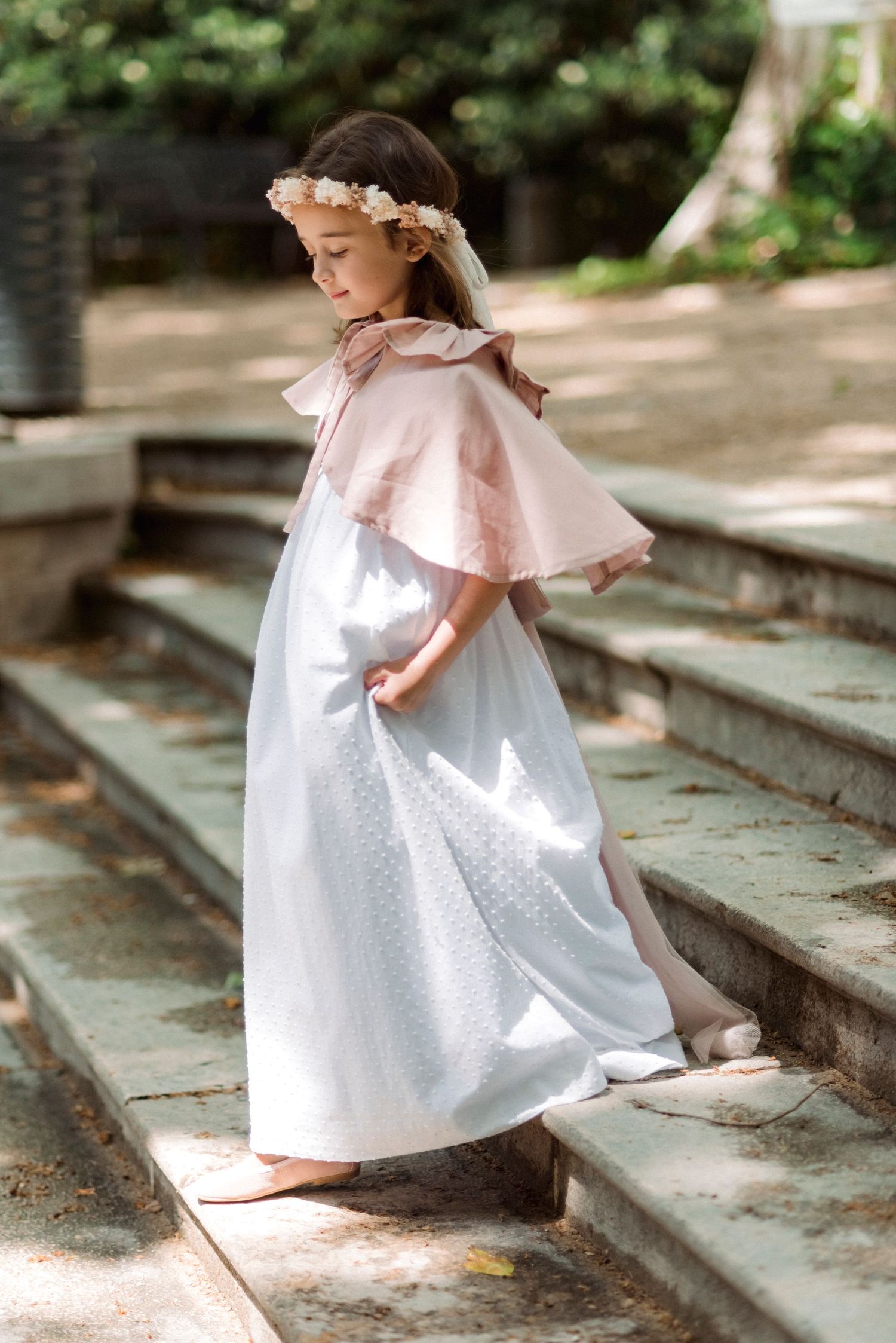 Young girl in white dotted dress and pink cape with flower crown walking up stone steps.