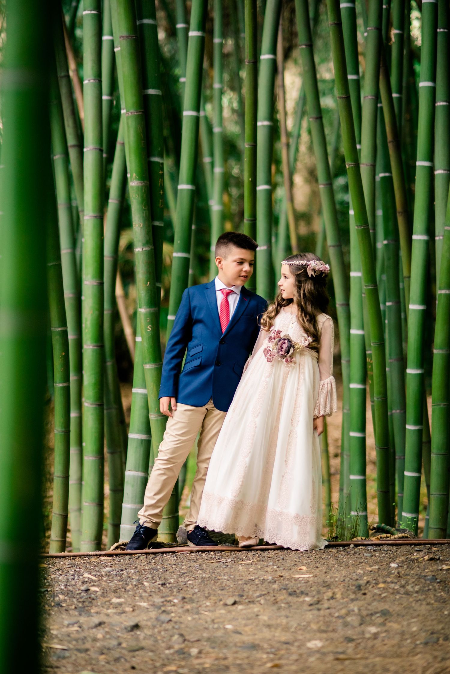 Boy in navy suit and girl in white dress with floral crown standing in bamboo forest.