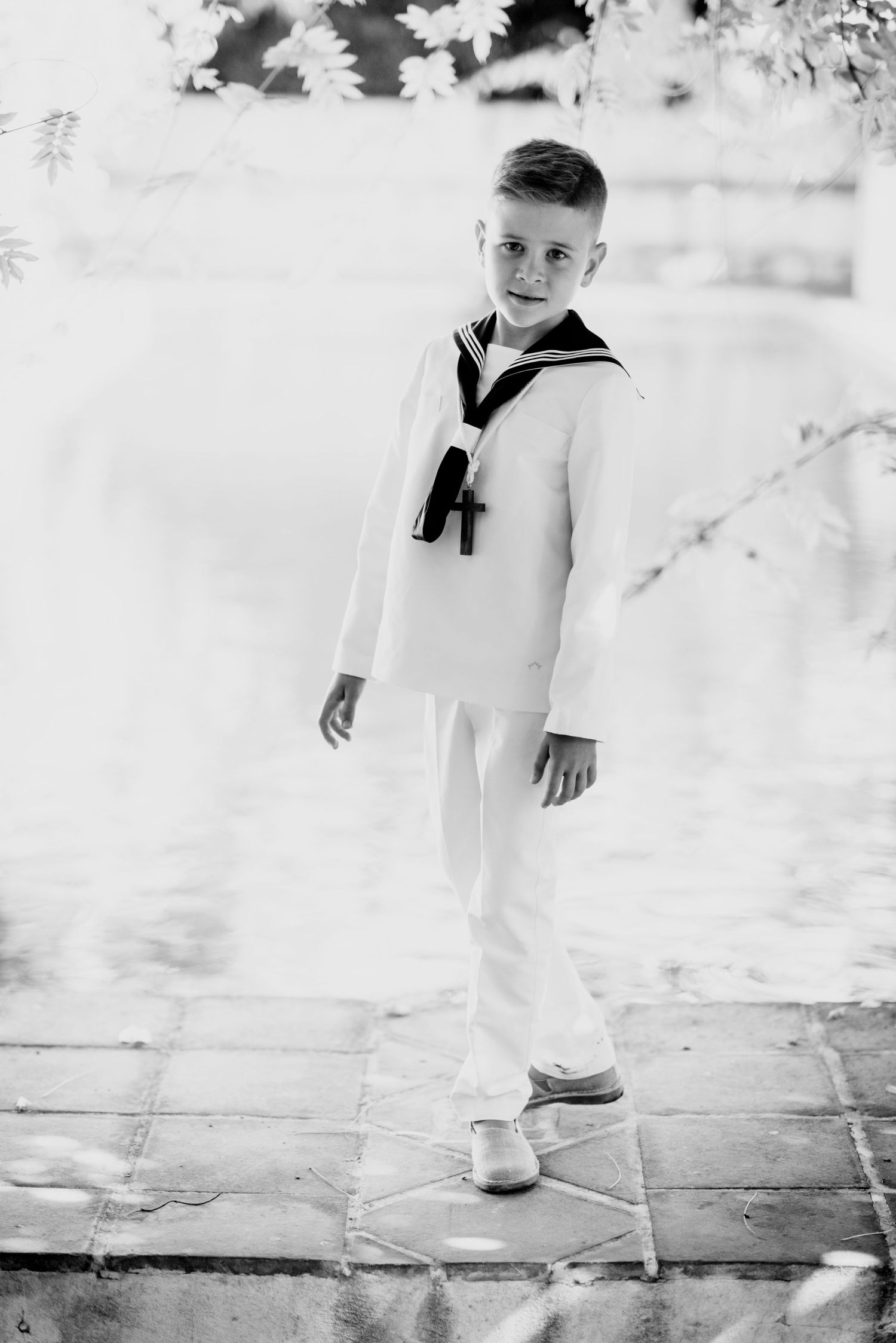 Black and white portrait of smiling boy in white sailor suit standing near reflective water.