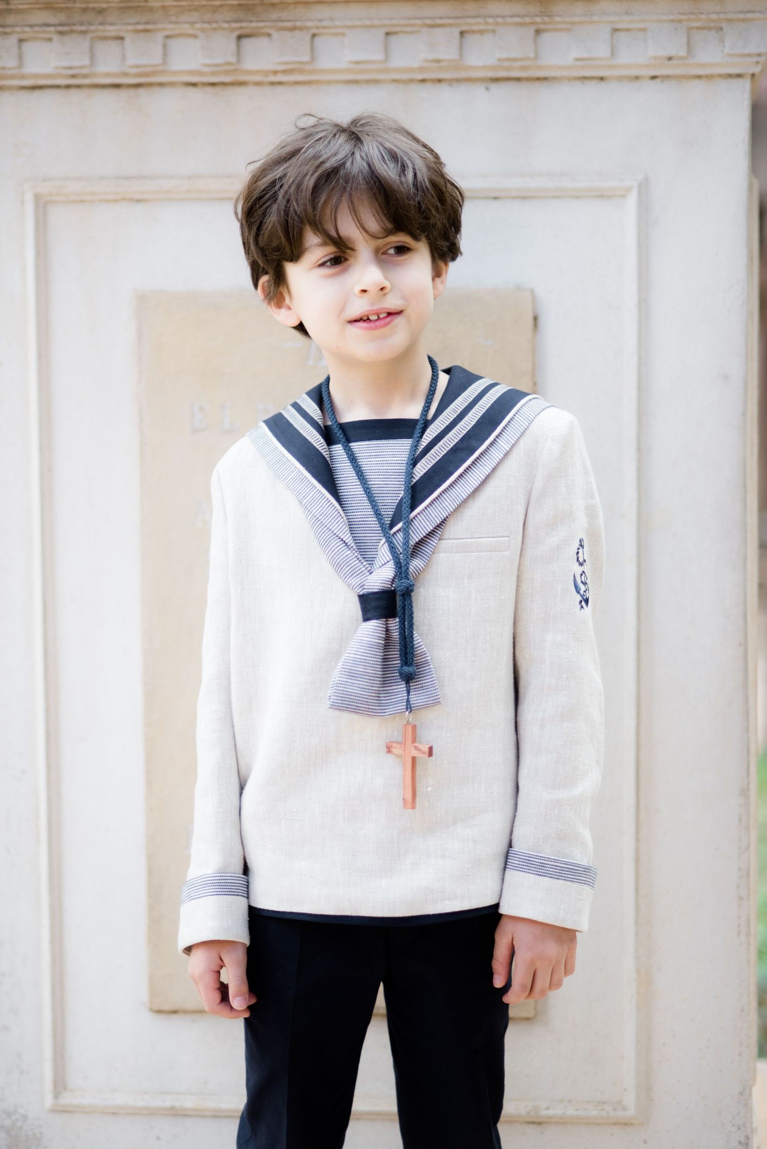 A young boy in a sailor communion outfit with a wooden cross necklace smiles warmly.