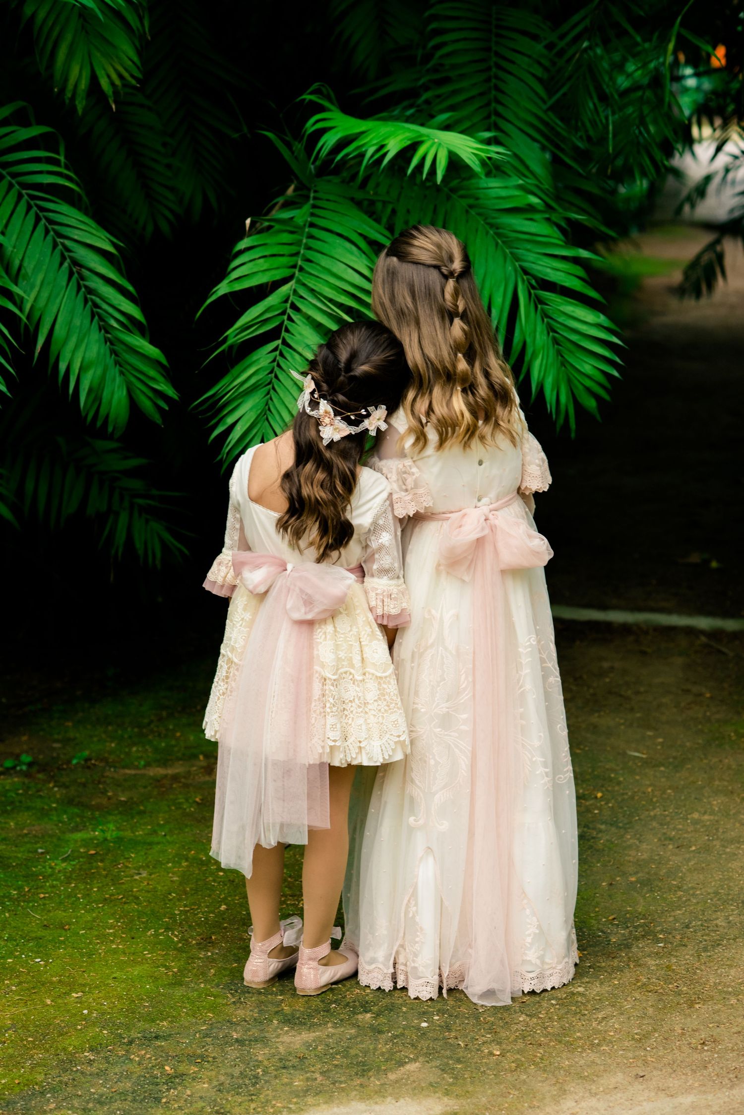 Two girls in pink and ivory dresses standing by palm fronds.