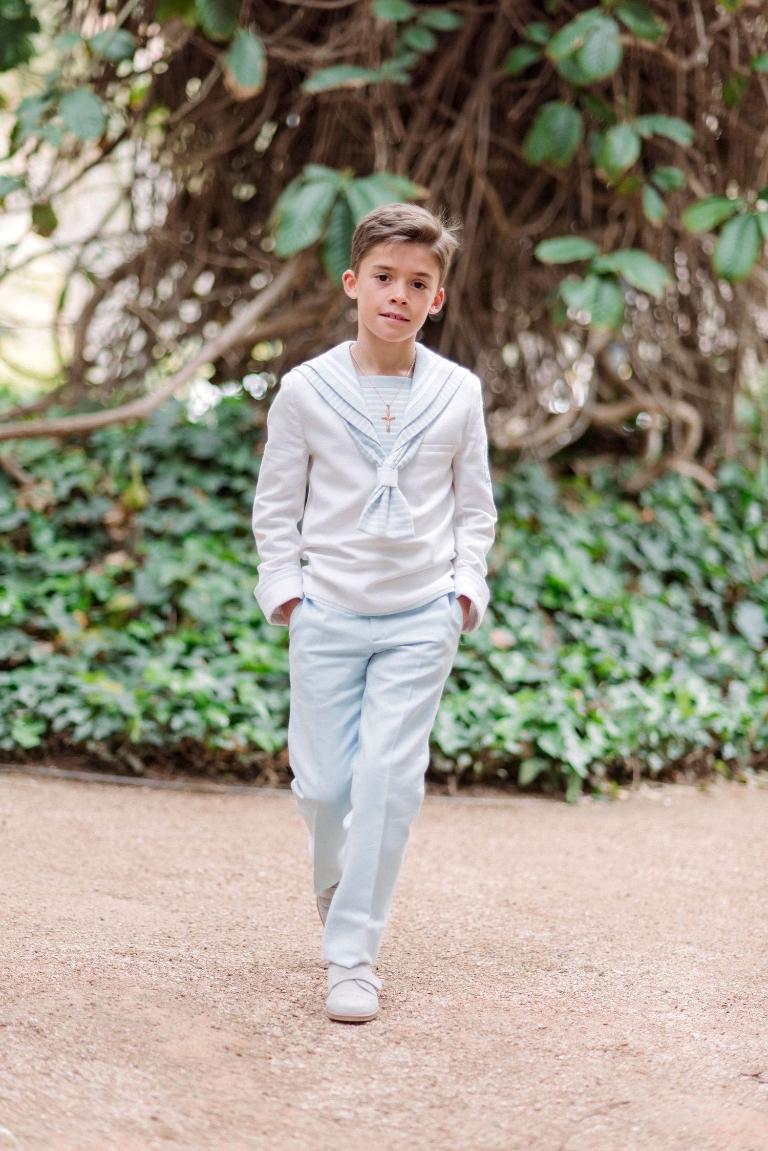 Boy in white sailor-style communion outfit walks along garden path with ivy-covered wall behind him.