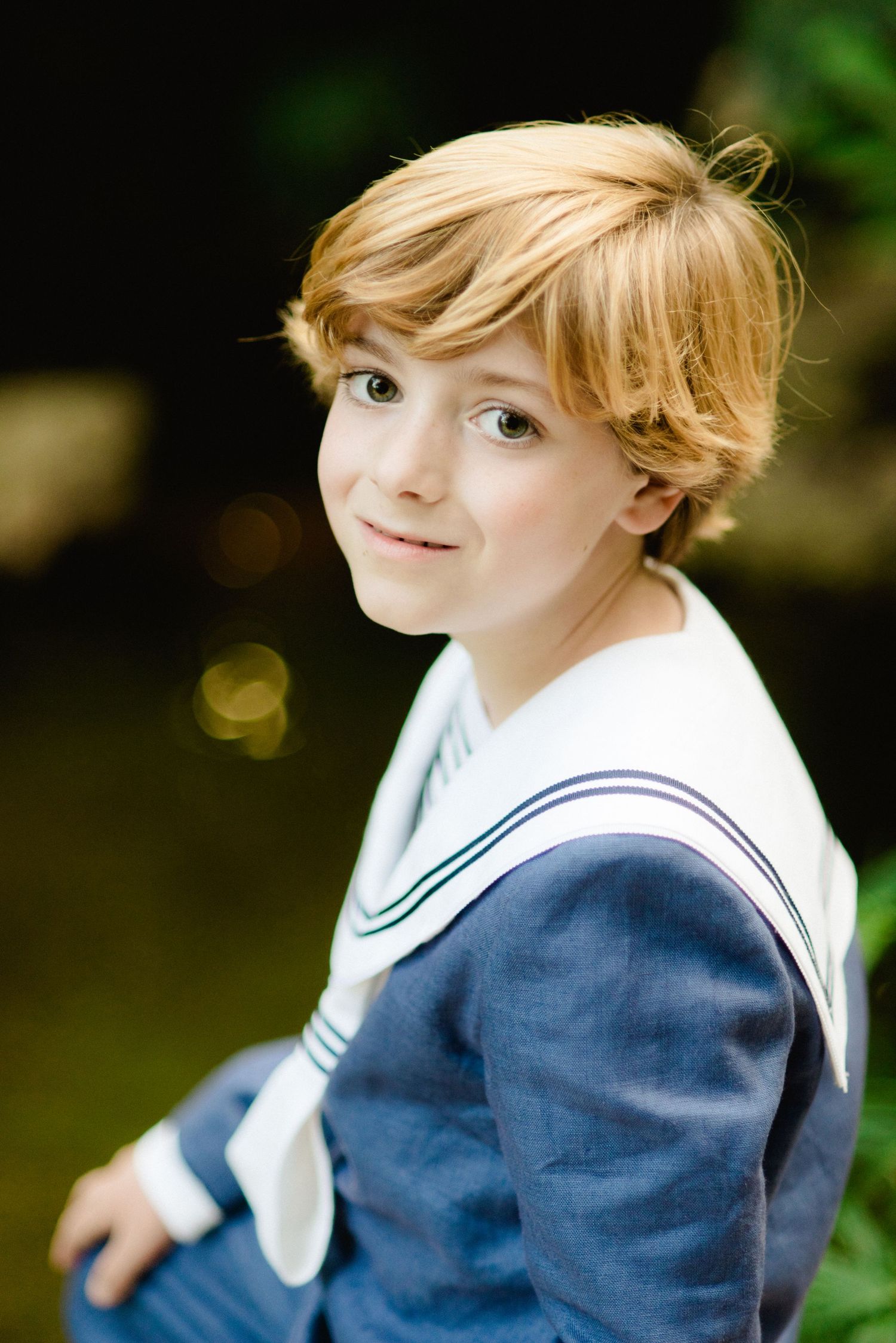 Close-up portrait of a blonde boy in navy sailor suit smiling in a garden.