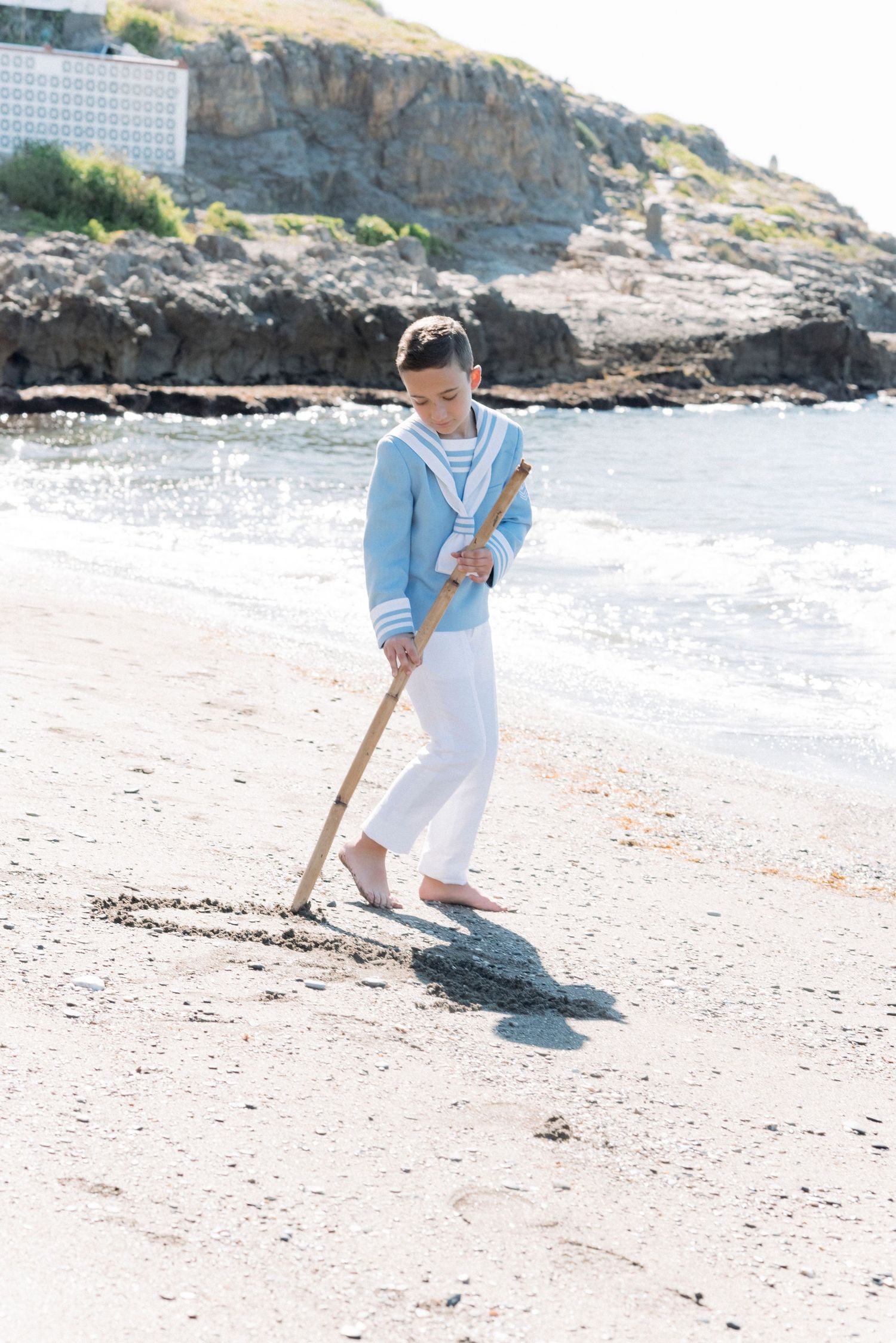 A young boy in a sailor outfit draws in the sand with a stick on a sunny beach.