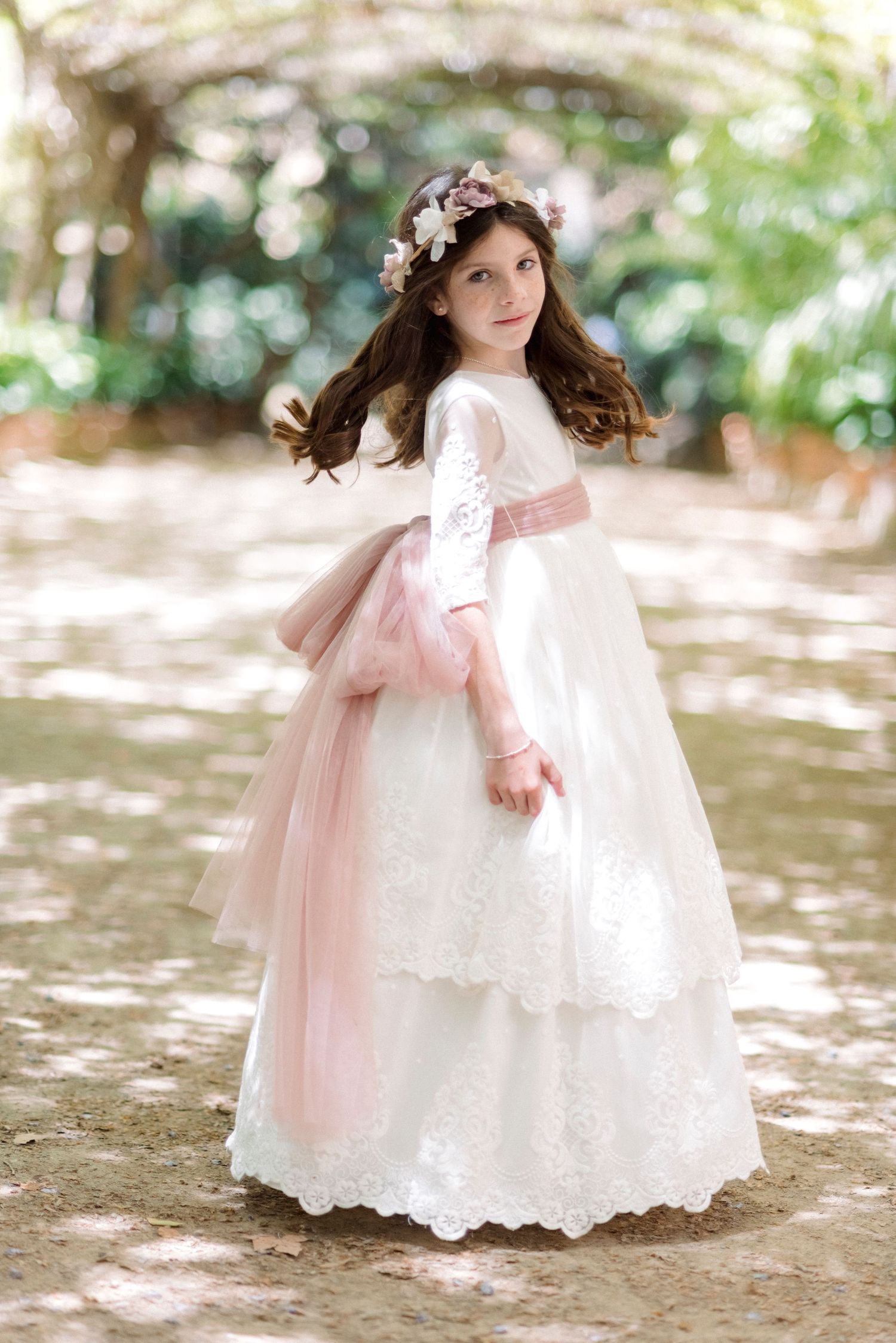 Young girl in white lace communion dress with pink tulle sash and floral crown in garden.