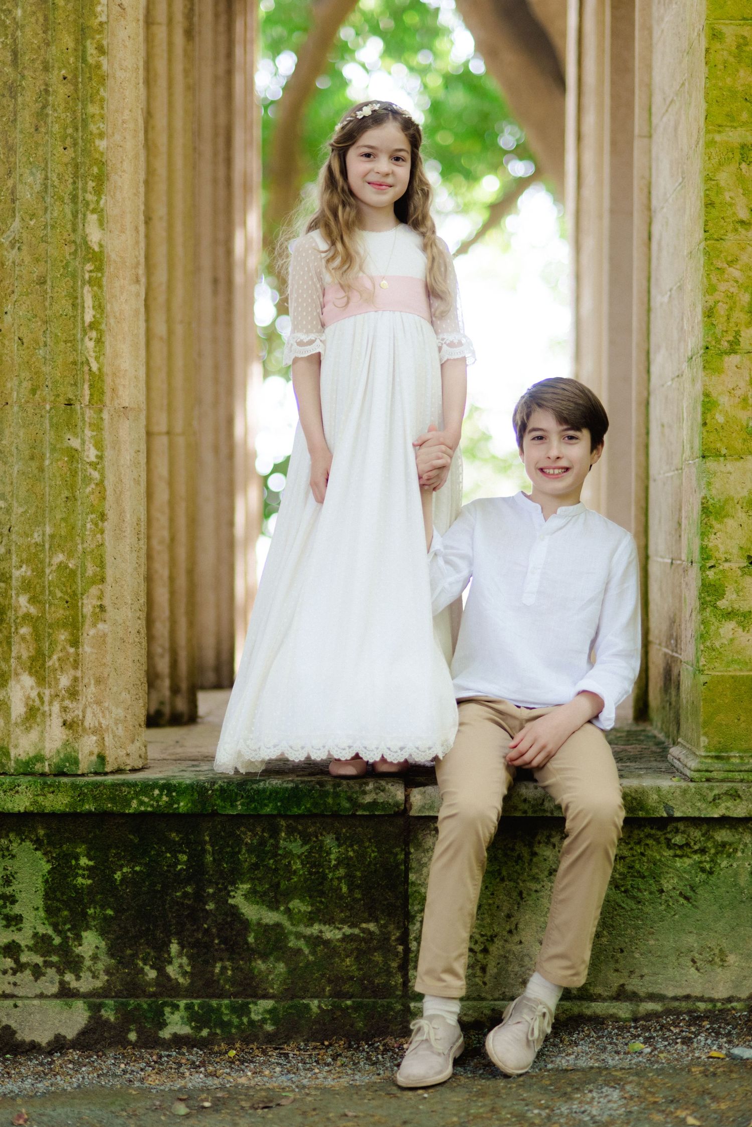 Girl in white communion dress and boy in white shirt posing under stone archway.
