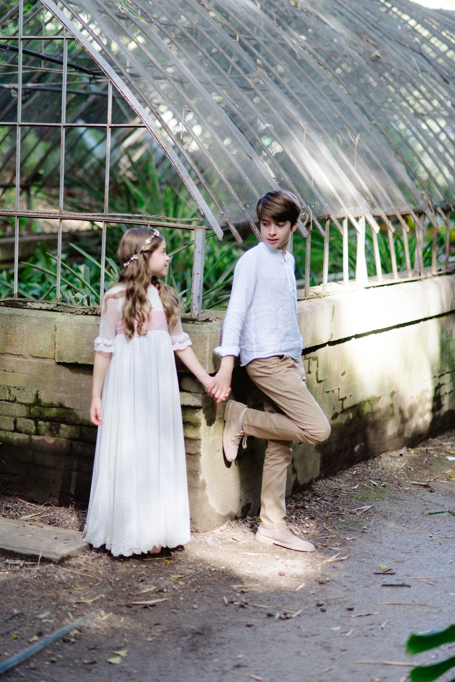 A boy and girl in elegant white communion outfits hold hands near a greenhouse wall.
