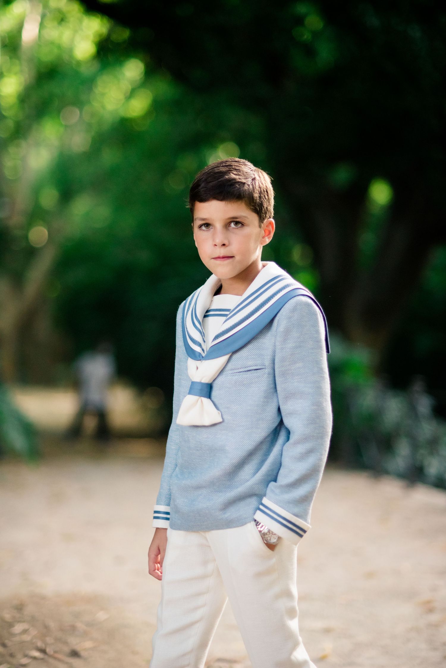 Young boy wearing light blue sailor collar sweater and white trousers posing outdoors in park.