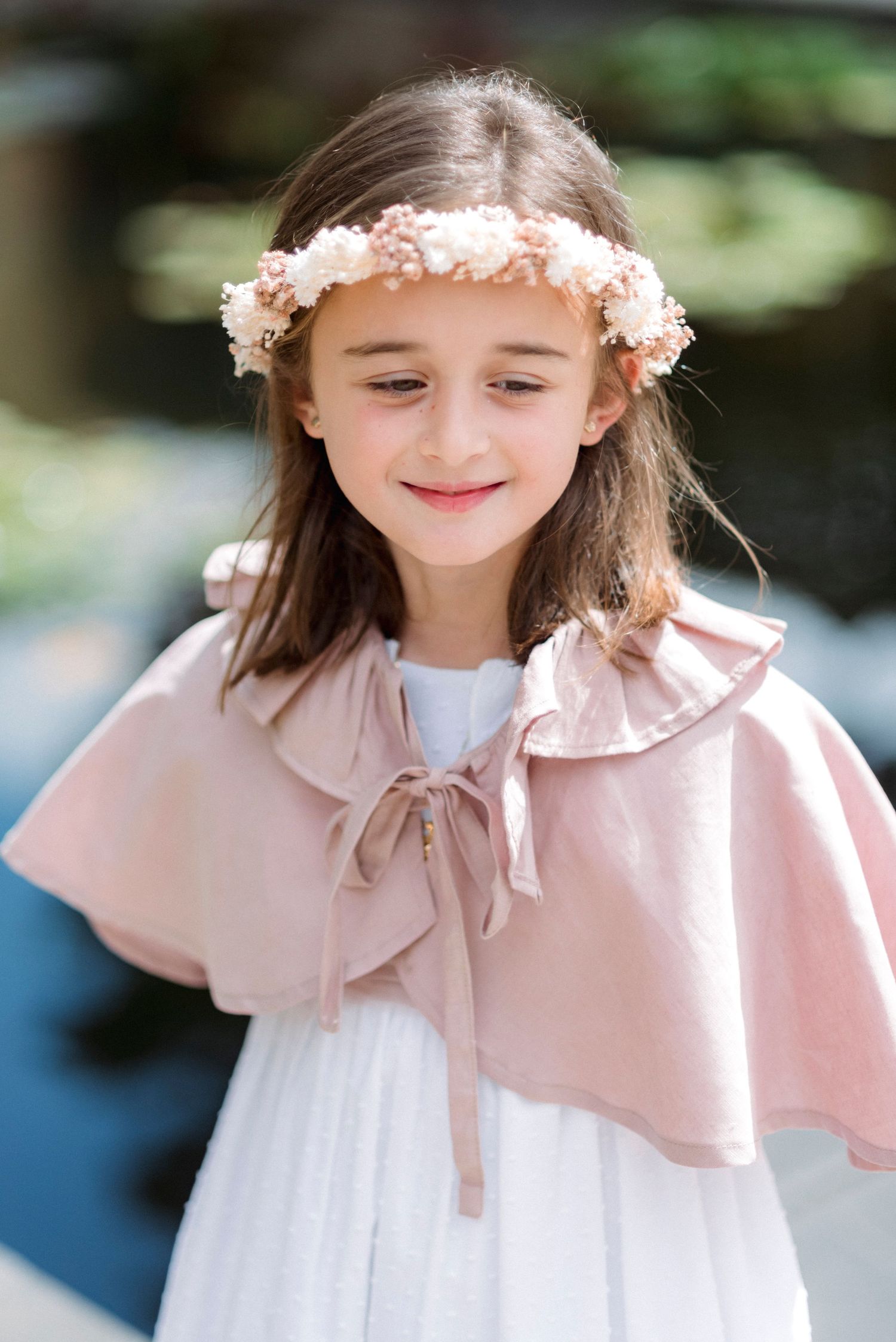 Young girl in white dress with pink ruffle cape and floral crown smiling softly outdoors.