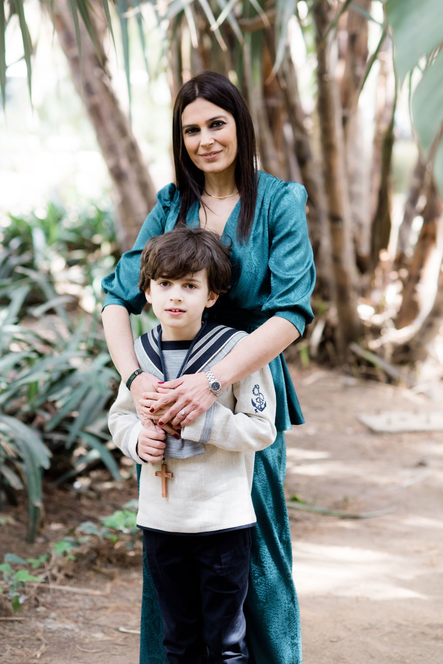 A dark-haired woman in teal embraces her young son holding a cross in a garden.