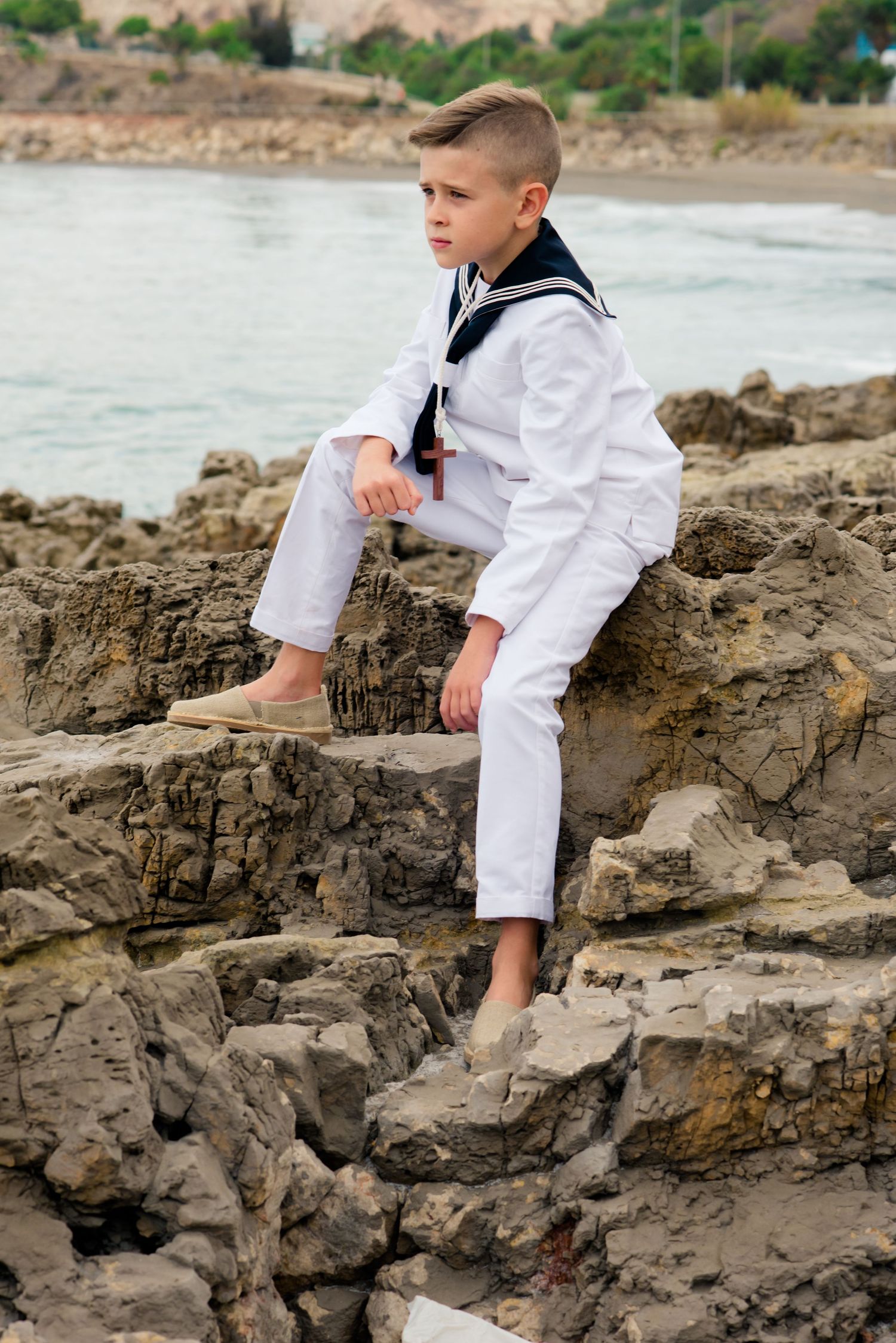 Boy in white sailor uniform sits barefoot on rugged coastal rocks with sea behind him.
