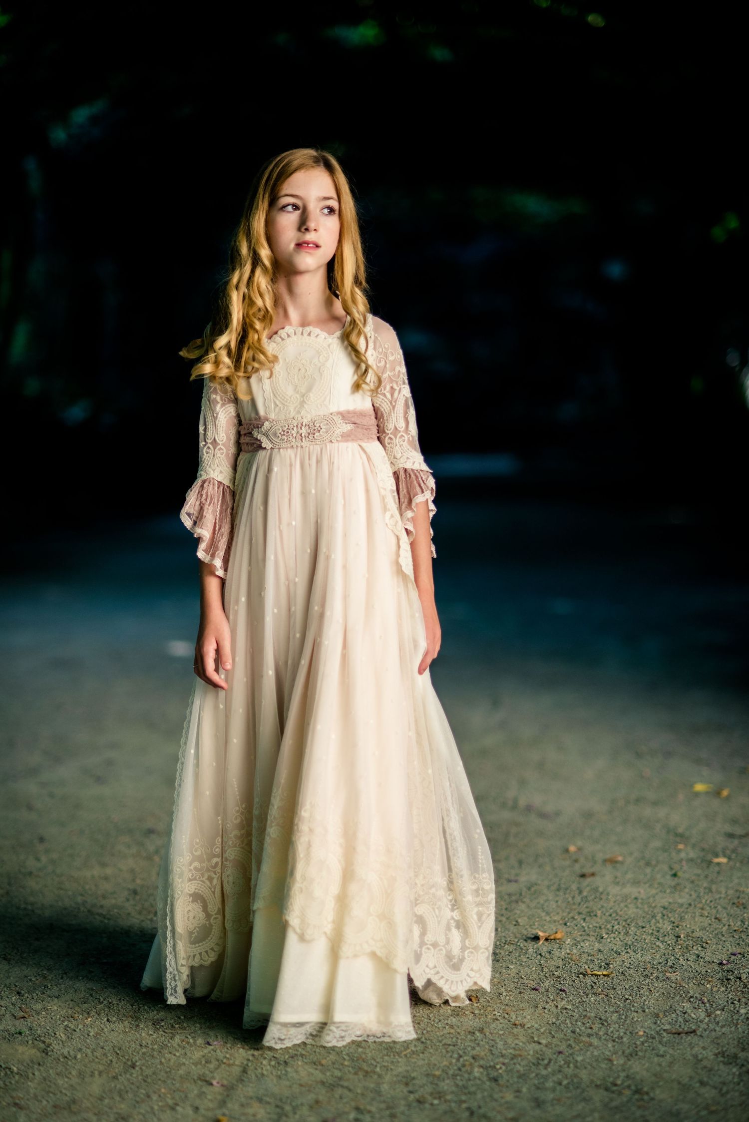 Young girl in elegant cream lace gown with bell sleeves poses in dramatic dark outdoor scene.