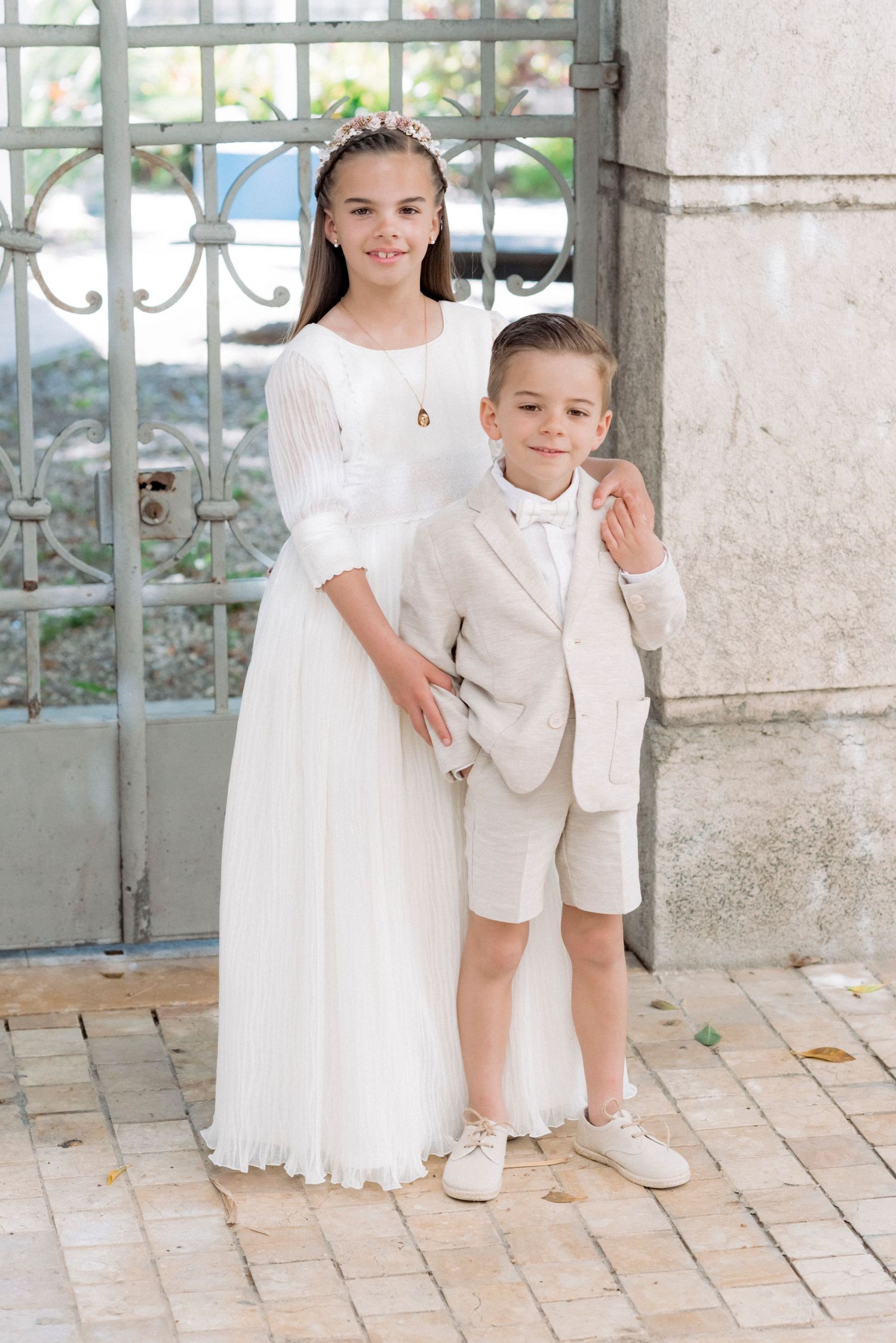 Girl in white communion dress and boy in beige suit posing by ornate iron gate.
