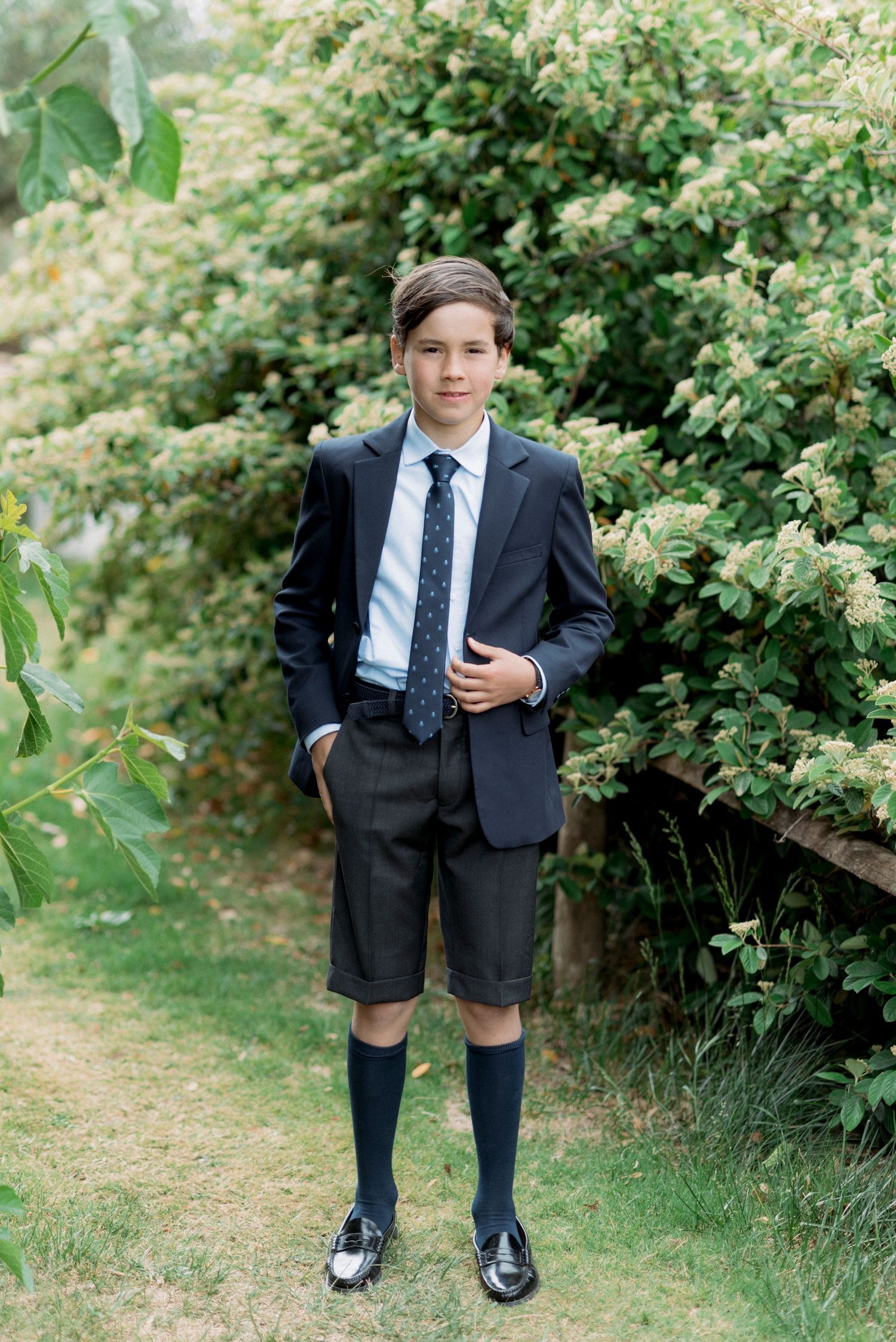 Boy in navy blazer, tie, shorts, and knee socks posing in front of flowering green bushes.