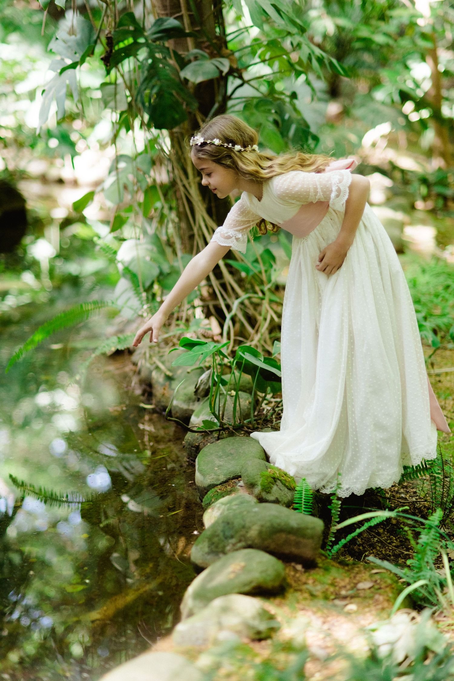 Girl in white lace dress with floral crown reaching toward a tranquil garden pond.