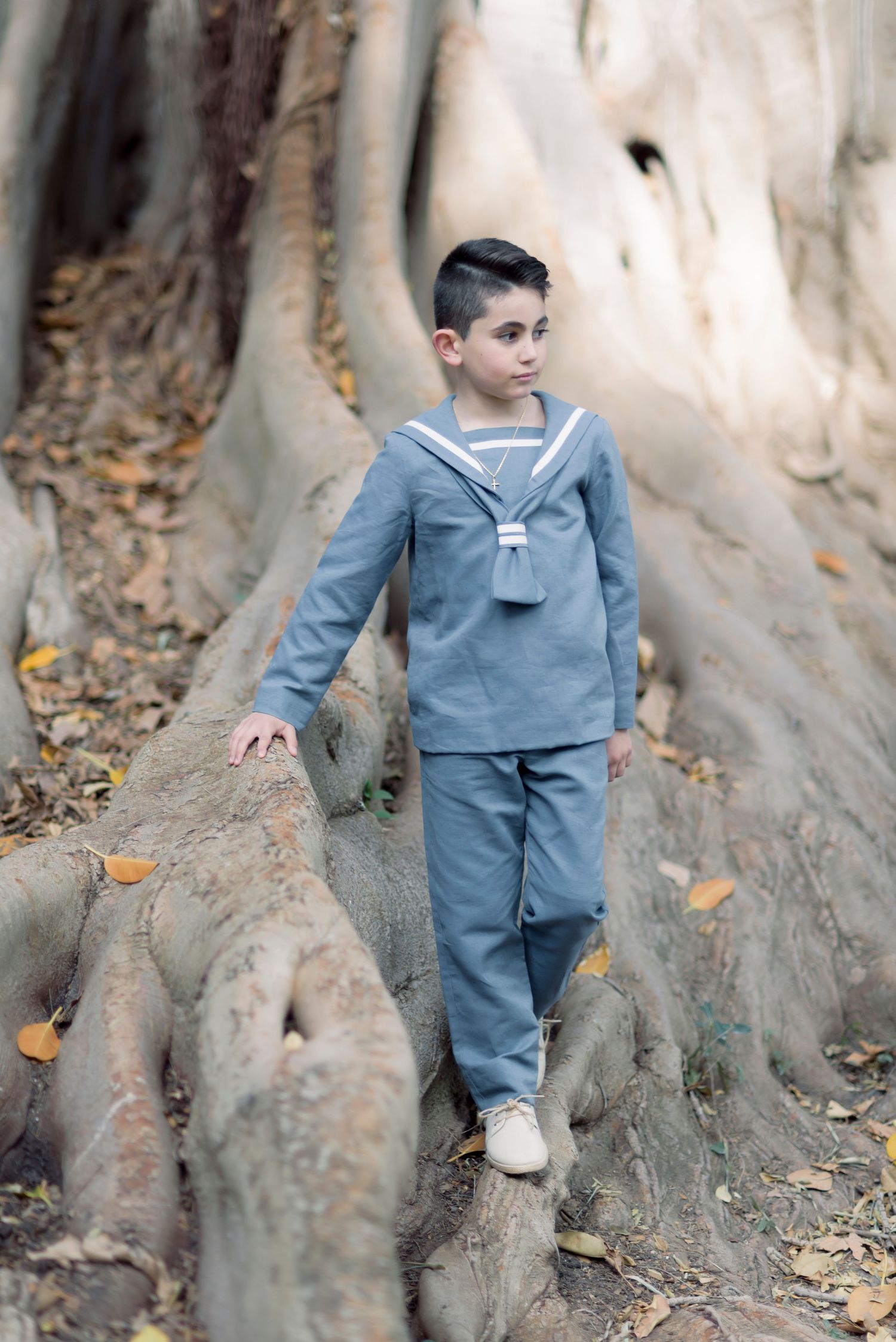 Boy in vintage gray sailor suit posing among large tree roots in autumn forest.