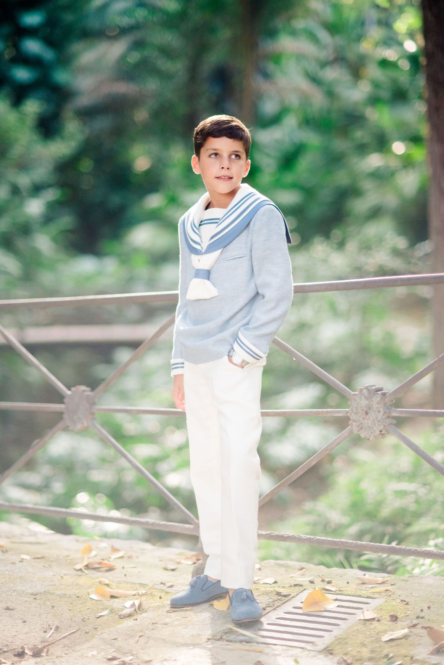 Boy in sailor-style blue sweater and white pants standing by metal fence in green park.
