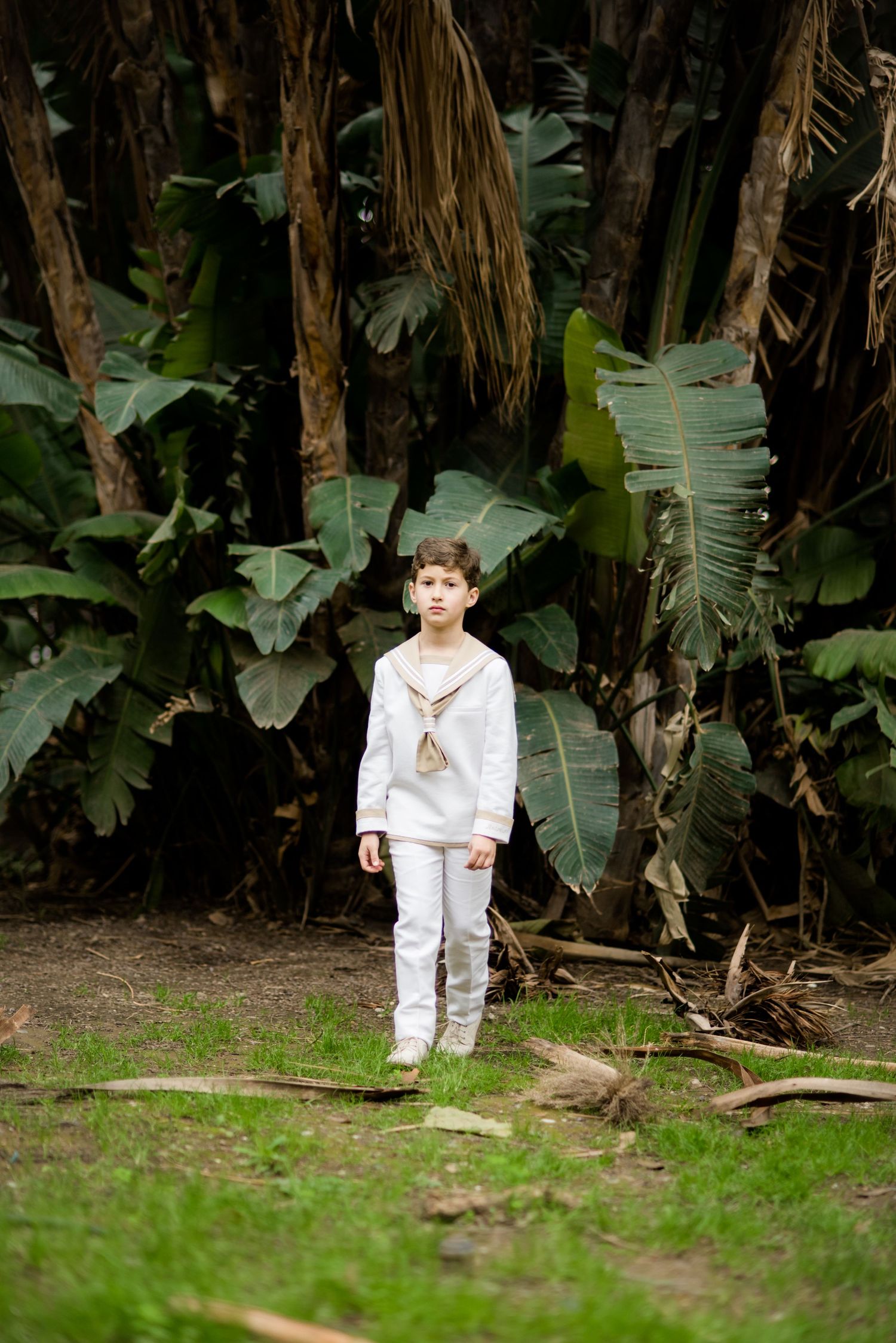 Boy in white sailor outfit standing in tropical garden.