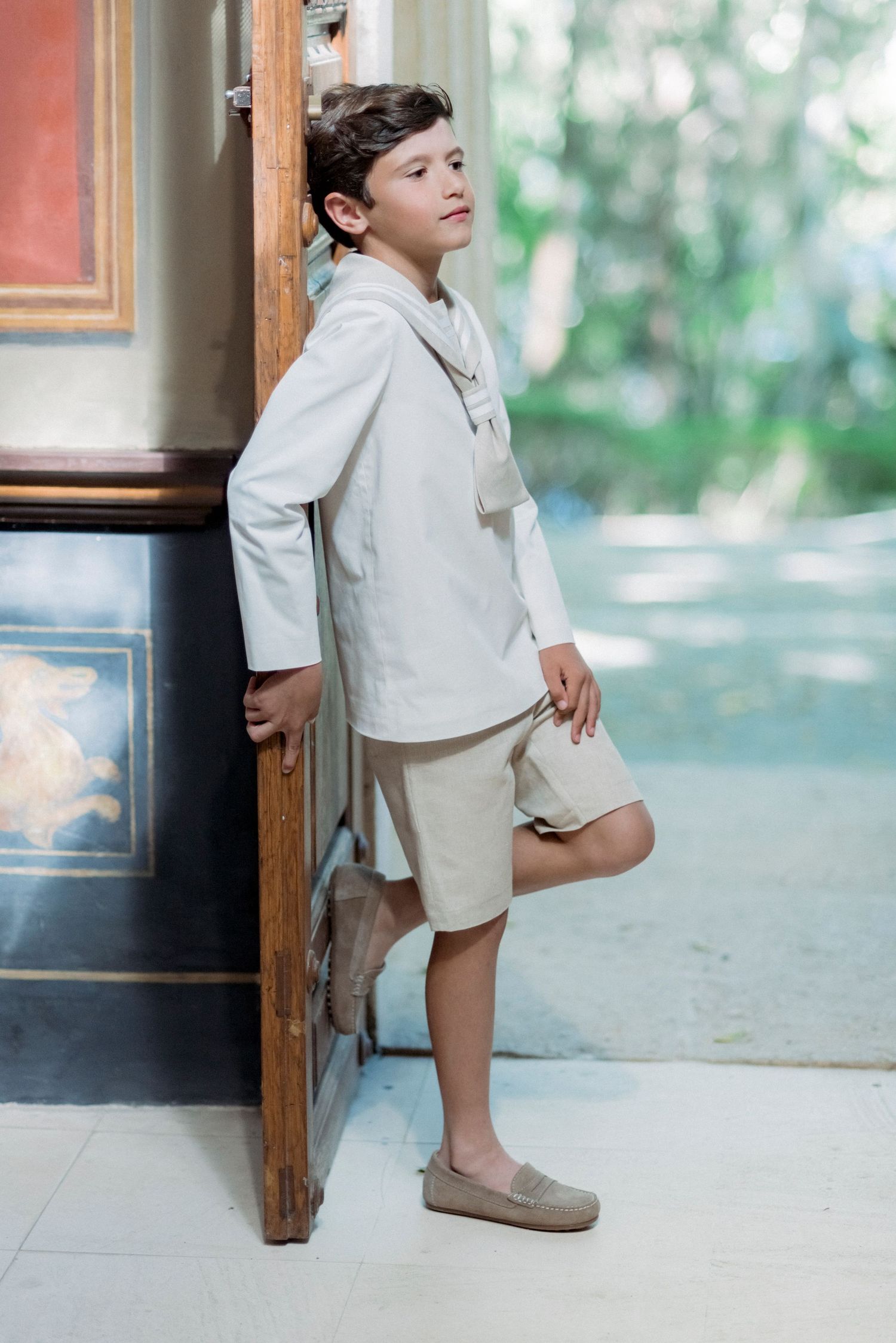 Boy in white sailor outfit leaning against ornate wooden door in elegant interior.