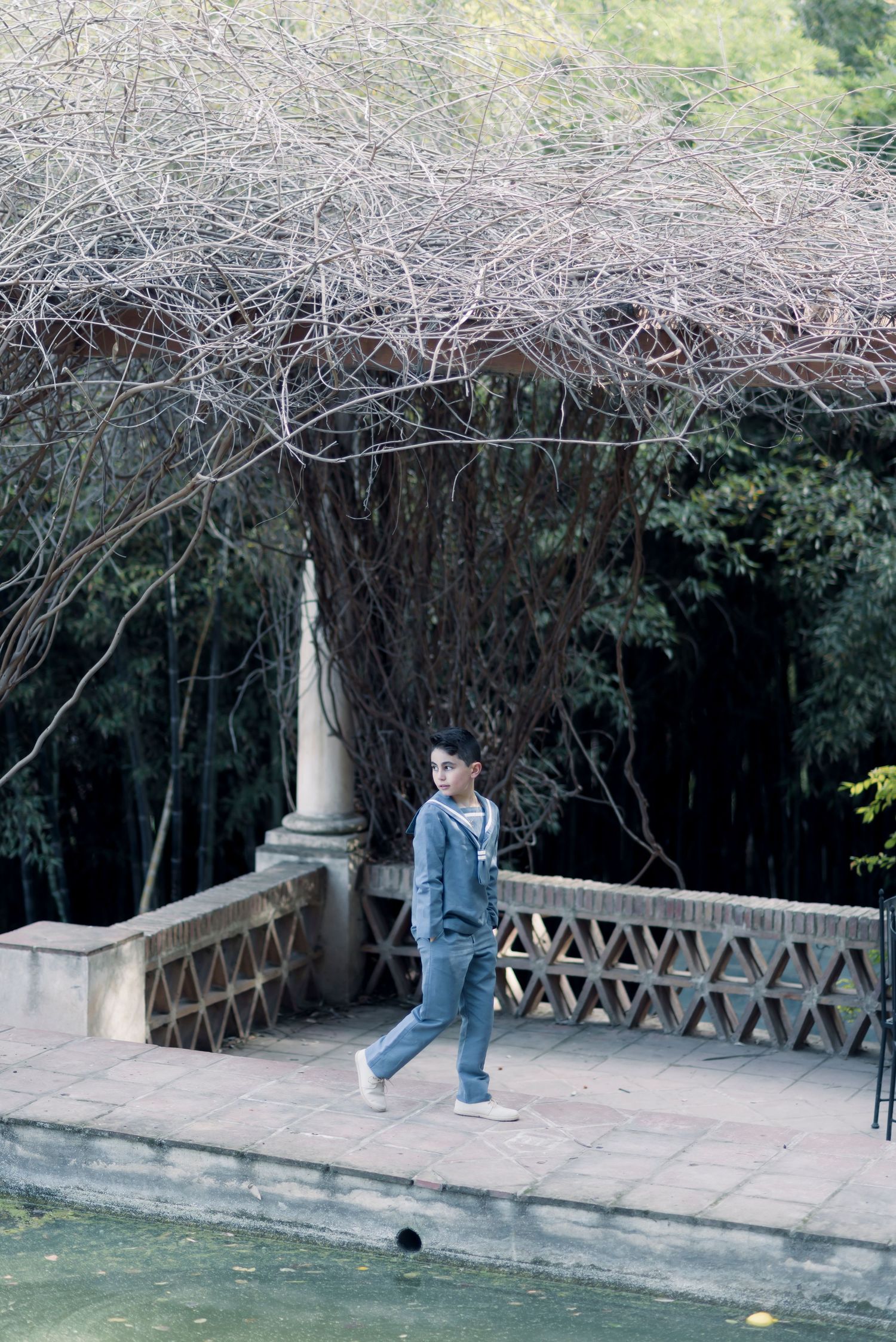 Young boy in gray sailor suit walking on a stone bridge surrounded by bare trees.