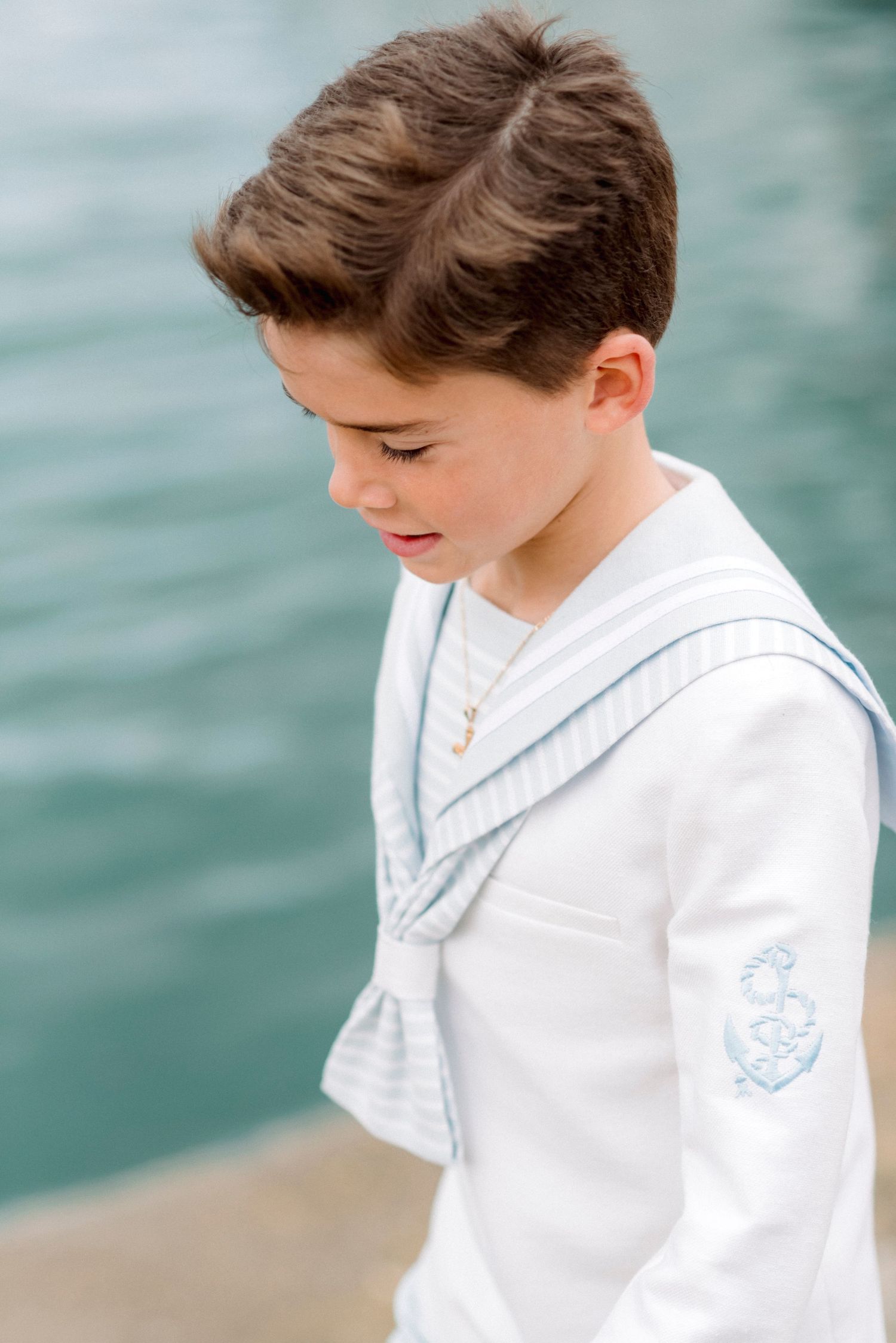 Young boy in white sailor suit with light blue trim poses near water for First Communion.